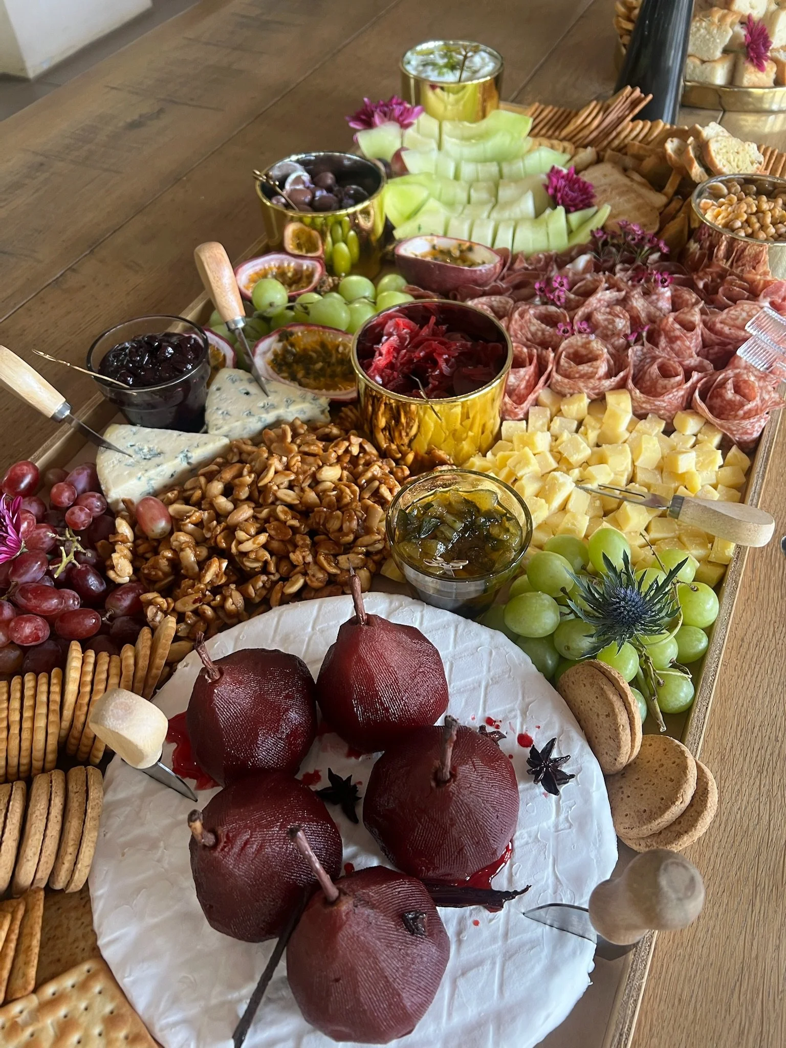 Assorted fruits, cheeses, crackers, nuts, and condiments arranged on a wooden table for a charcuterie board display.