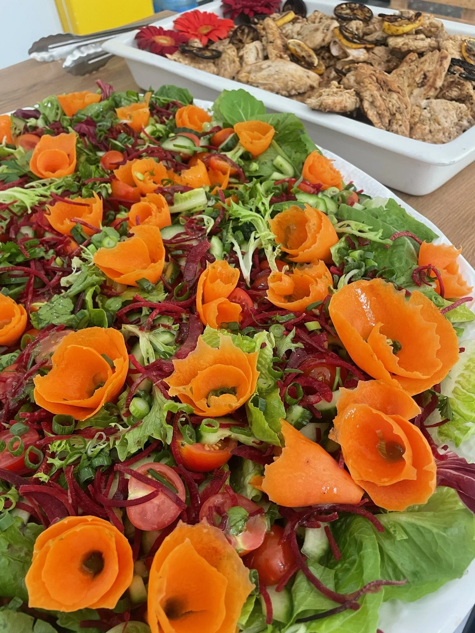 A colorful salad made with leafy greens, cherry tomatoes, shredded red cabbage, chopped cucumbers, and rolled slices of orange bell pepper, topped with sliced green onions, on a white platter. In the background, there is a tray of grilled chicken win