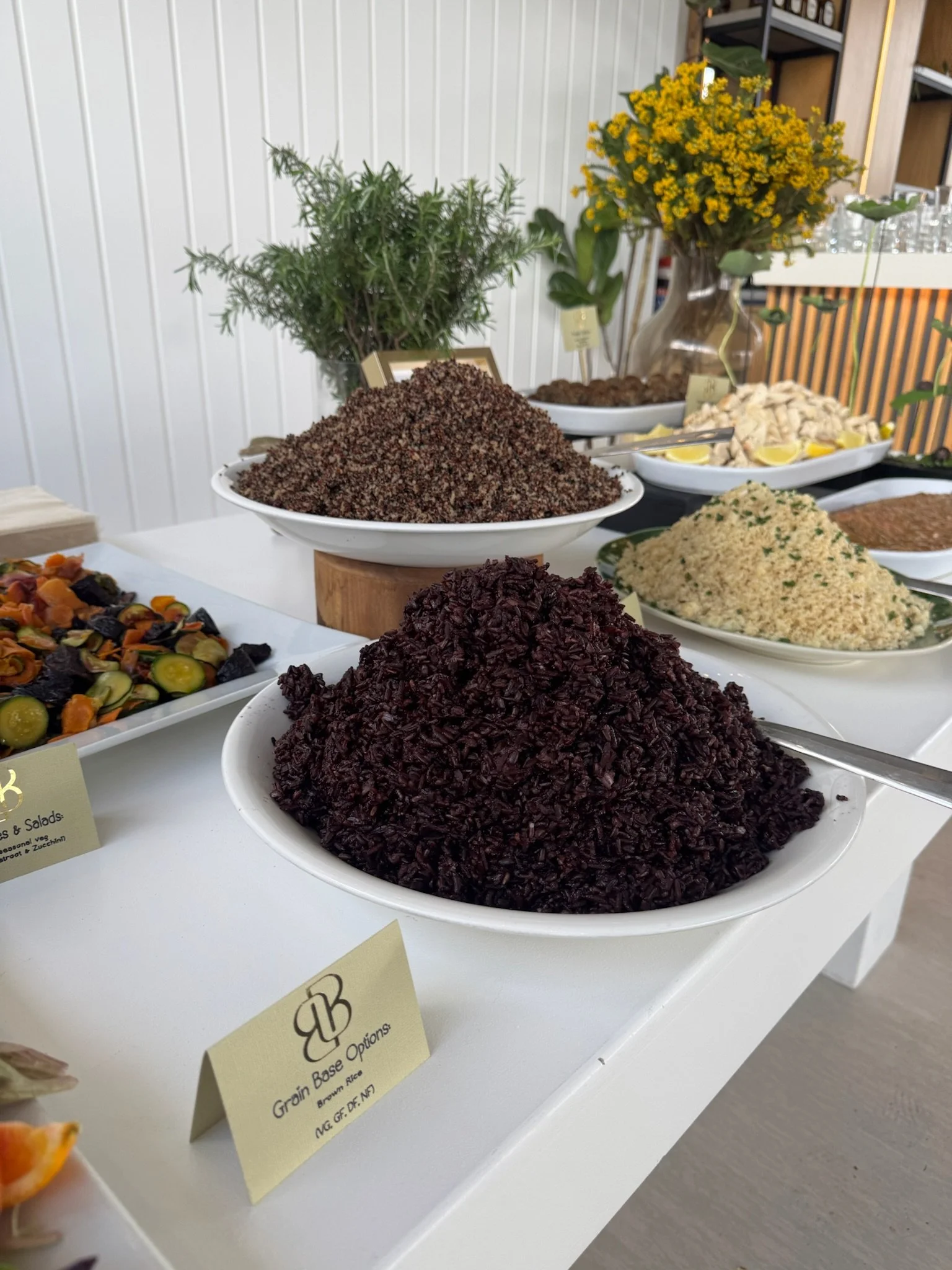 Bowls of cooked grains including black rice, quinoa, and seasoned rice on a white table with flowers and leaves in the background.