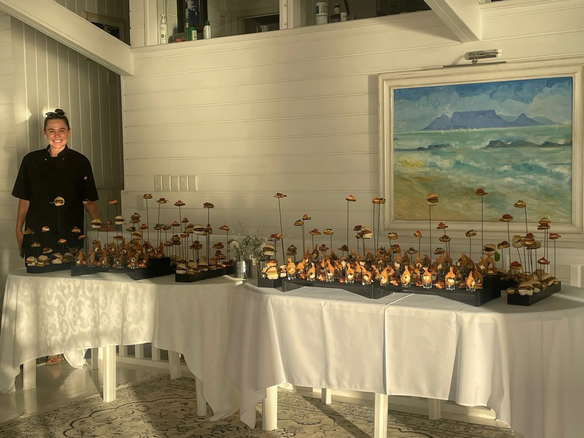 A woman in a black uniform smiling standing behind a table with an array of small sandwiches or appetizers on black trays, set in a room with white paneled walls and a framed painting of a seascape hanging on the wall.