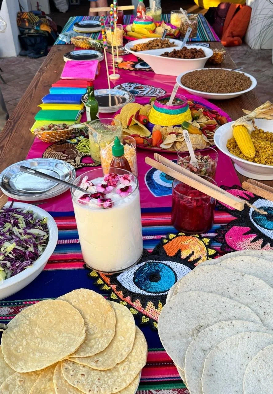 Colorful Mexican-themed party table with tacos, corn, salads, rice, and beverages, decorated with vibrant textiles and festive decorations.