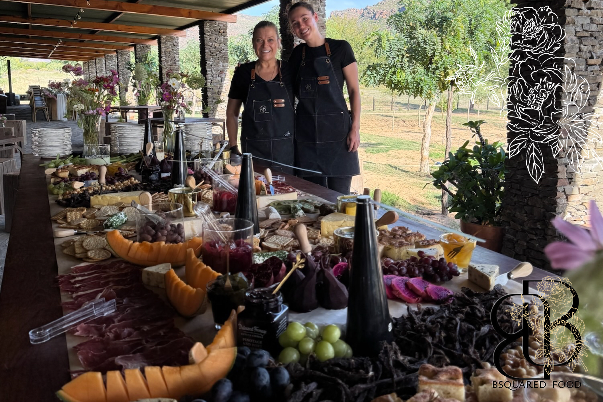 Two women wearing black aprons standing behind a table filled with various cheeses, fruits, and charcuterie at an outdoor event with trees and mountains in the background.