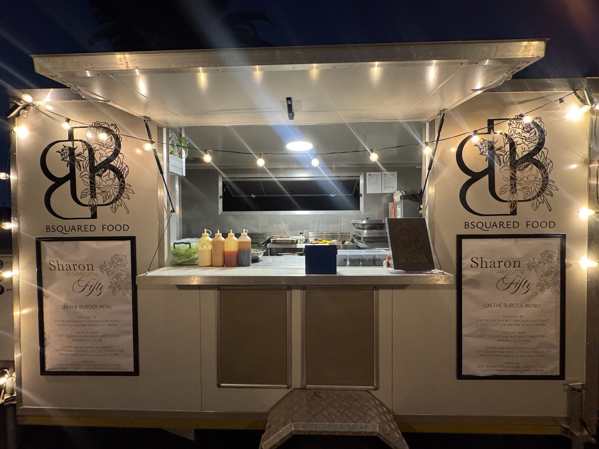 A small food stand with string lights and two signs reading 'Sharon is celebrating Fifty on the burger menu' with a list of burger ingredients. The stand has condiment bottles, a small menu board, and a kitchen area in the back.