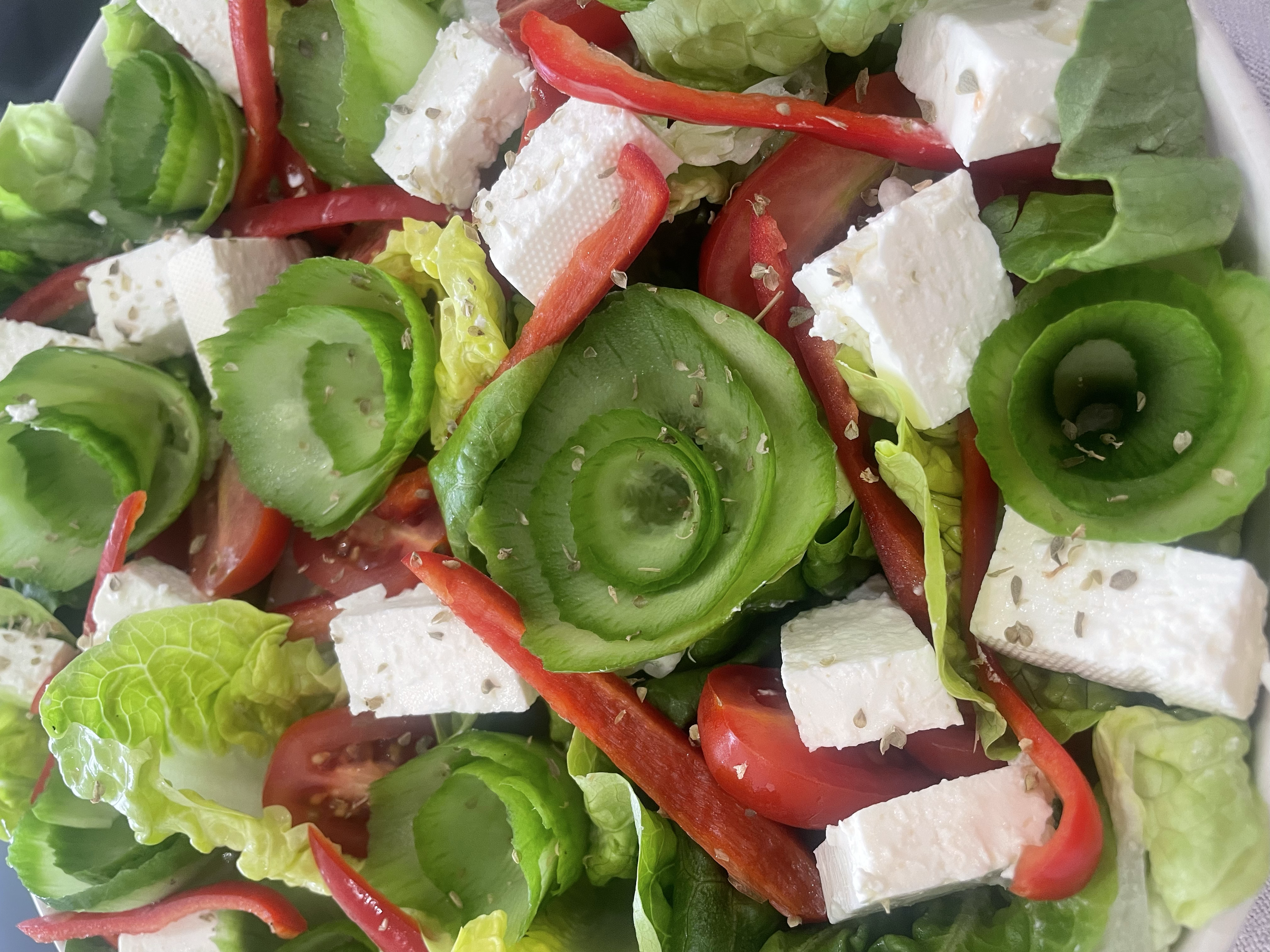 Close-up of fresh vegetable salad containing sliced cucumbers, cherry tomatoes, red bell peppers, lettuce, and cubes of feta cheese, sprinkled with herbs.