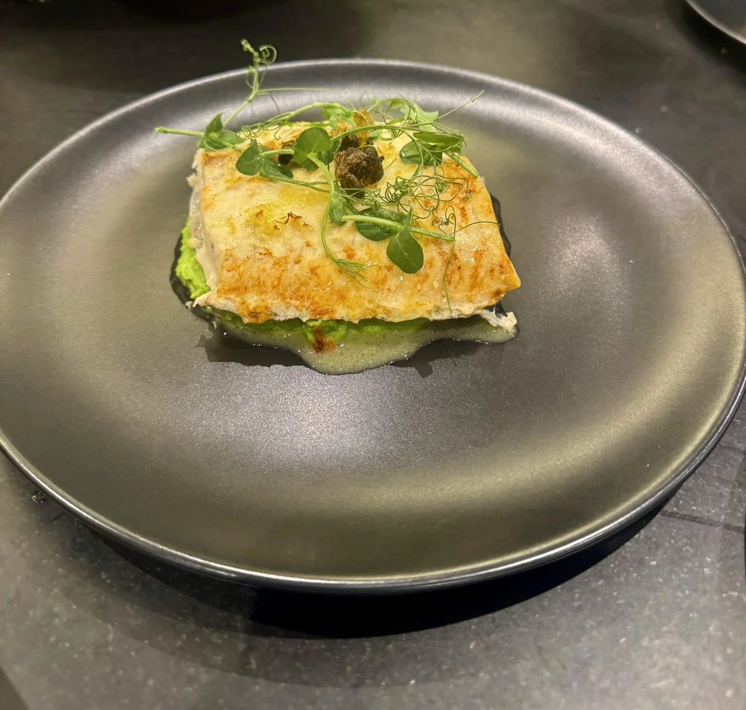 A plated dish featuring a piece of fried fish garnished with microgreens, served on a matte black plate.