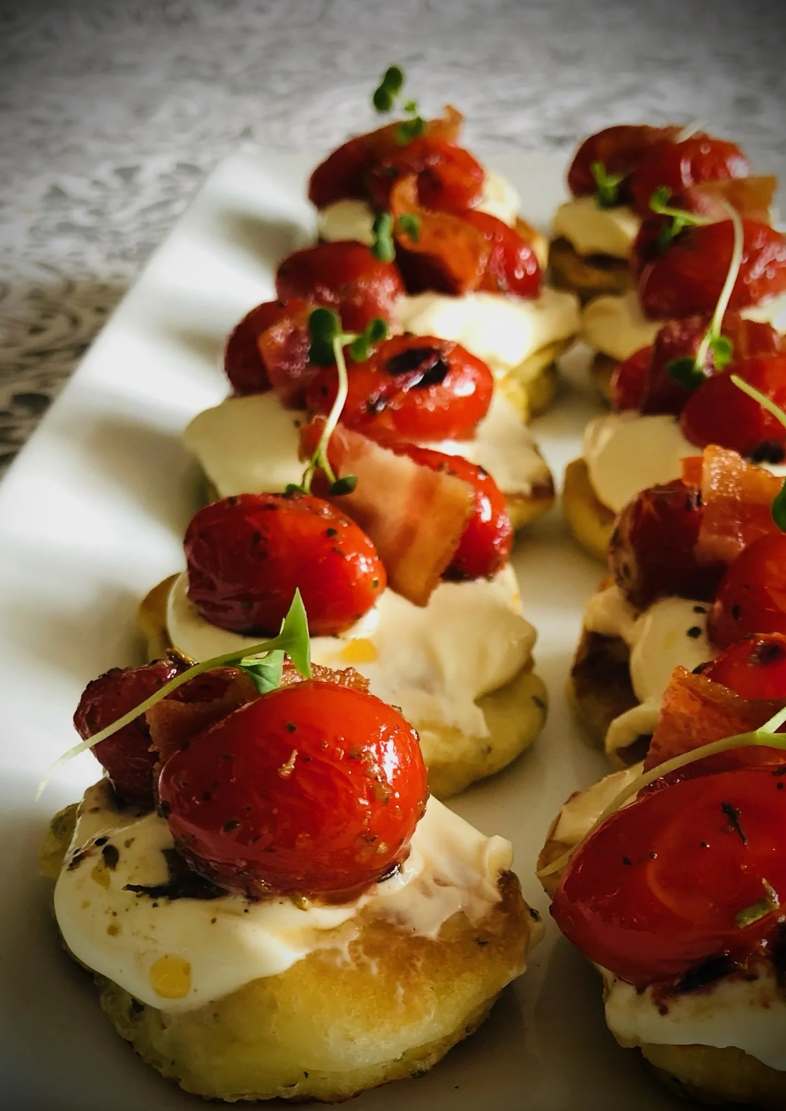 Close-up of miniature appetizers on a white plate, consisting of toasted bread topped with mayonnaise, cherry tomatoes, small bacon pieces, and microgreens.