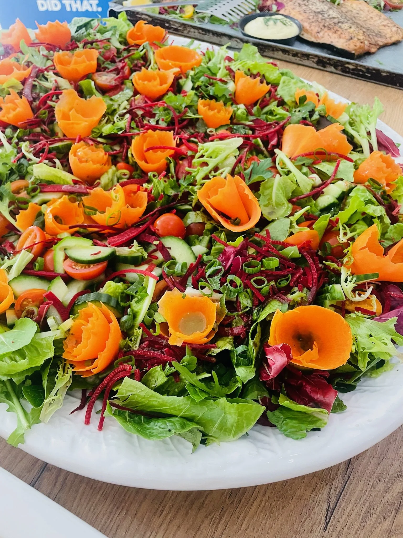 Mixed greens salad topped with carved orange carrots, cherry tomatoes, cucumbers, and chopped green onions, served on a white platter with a side of dipping sauce in the background.