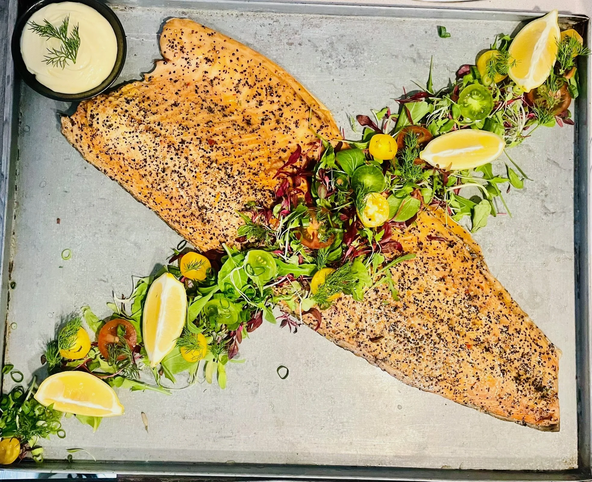 A tray with two perfectly cooked, seasoned salmon fillets garnished with fresh herbs and lemon wedges, accompanied by a colorful side salad with mixed greens, cherry tomatoes, and microgreens, and a small bowl of creamy dipping sauce.