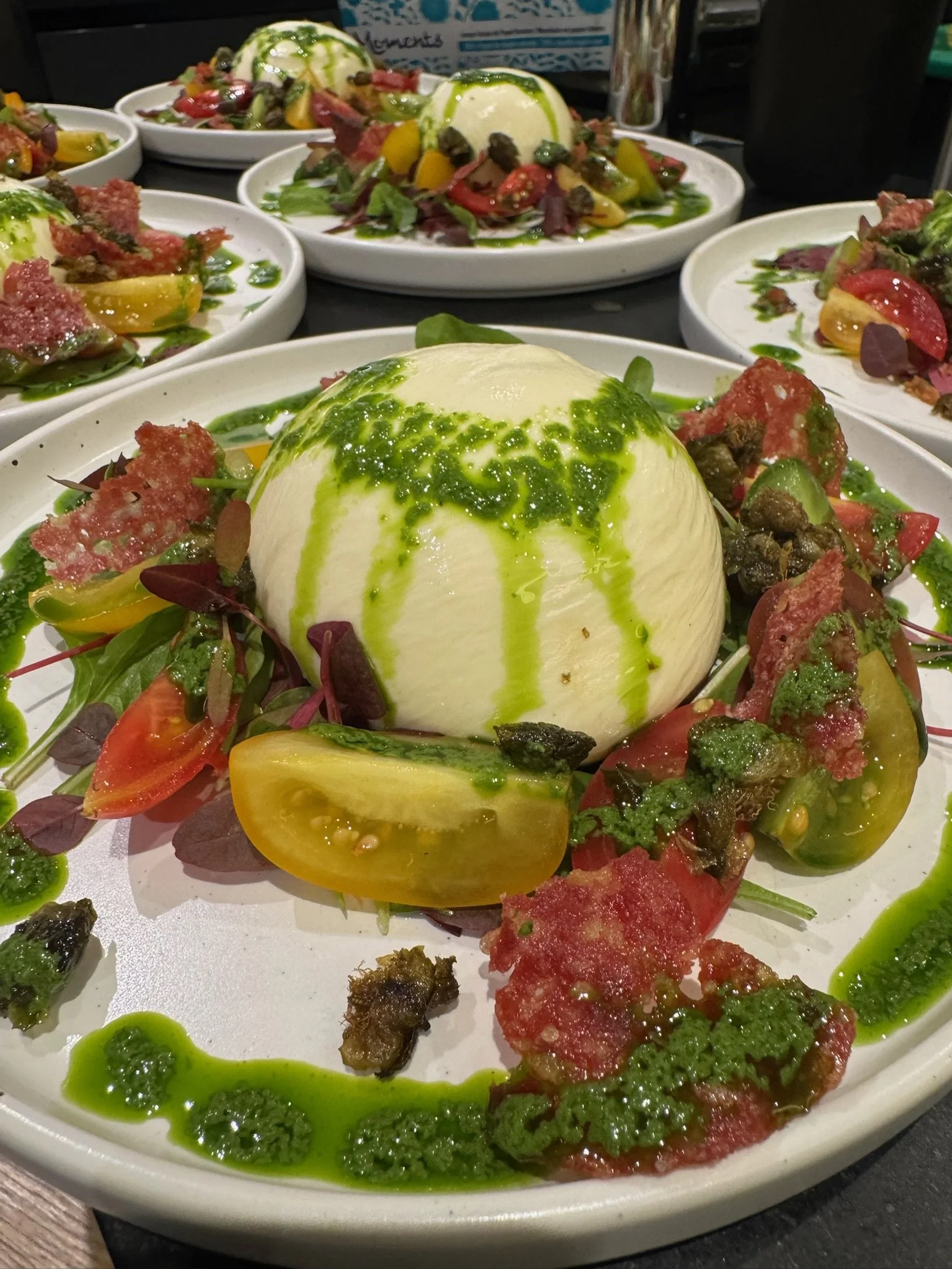 A Caprese salad with fresh mozzarella, heirloom tomatoes, basil pesto, and microgreens, garnished with a drizzle of green herb sauce and balsamic vinegar, served on a white plate.