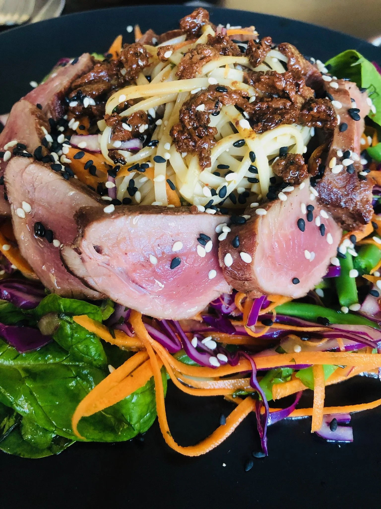 A plate of salad with sliced beef, shredded carrots, purple cabbage, and spinach, topped with noodles, and sesame seeds.