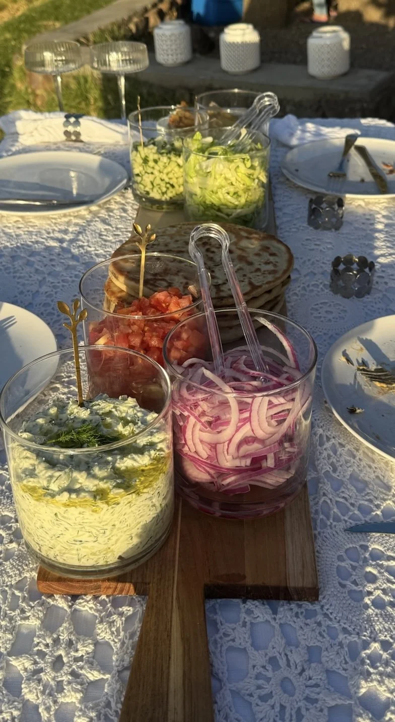 A table set with various bowls of chopped vegetables, a stack of flatbreads, and salad dressings on a white lace tablecloth at an outdoor gathering.