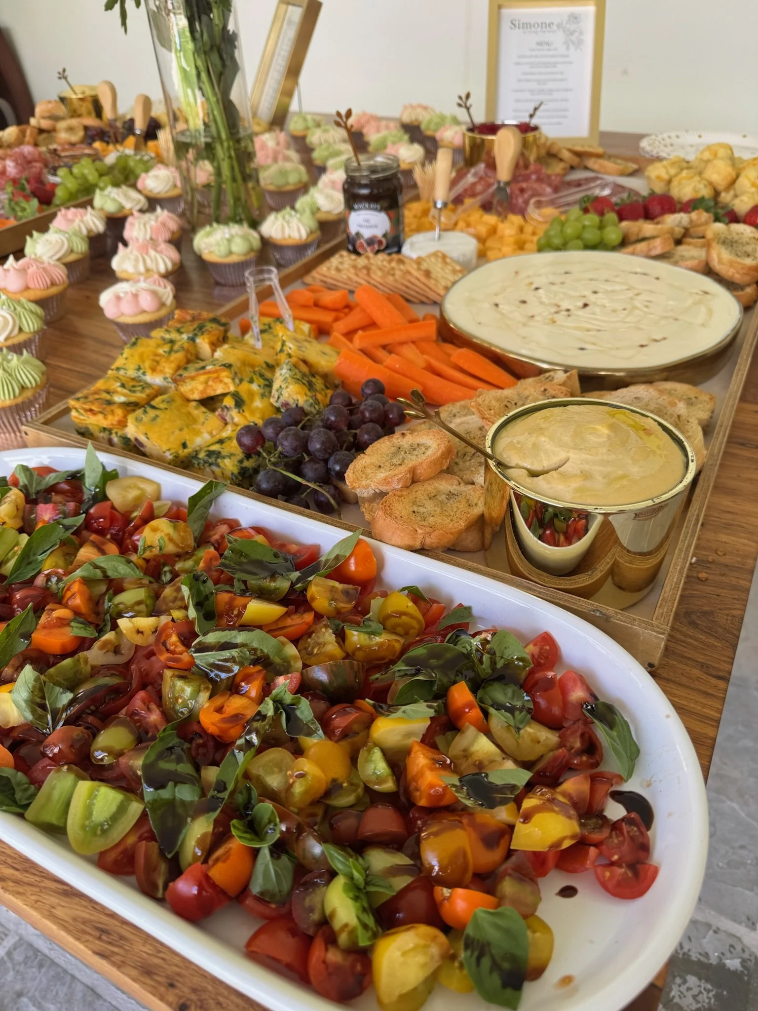 Assorted appetizers and desserts including cherry tomato salad, vegetable platter, cheese, crackers, grapes, cupcakes, bread, and dips set on a wooden table.