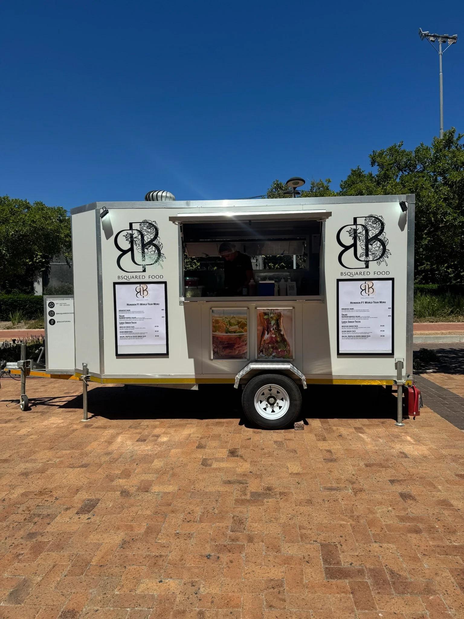 Food truck with a white exterior, black logos, and menu boards on each side, parked on brick pavement under a clear blue sky with some trees in the background.
