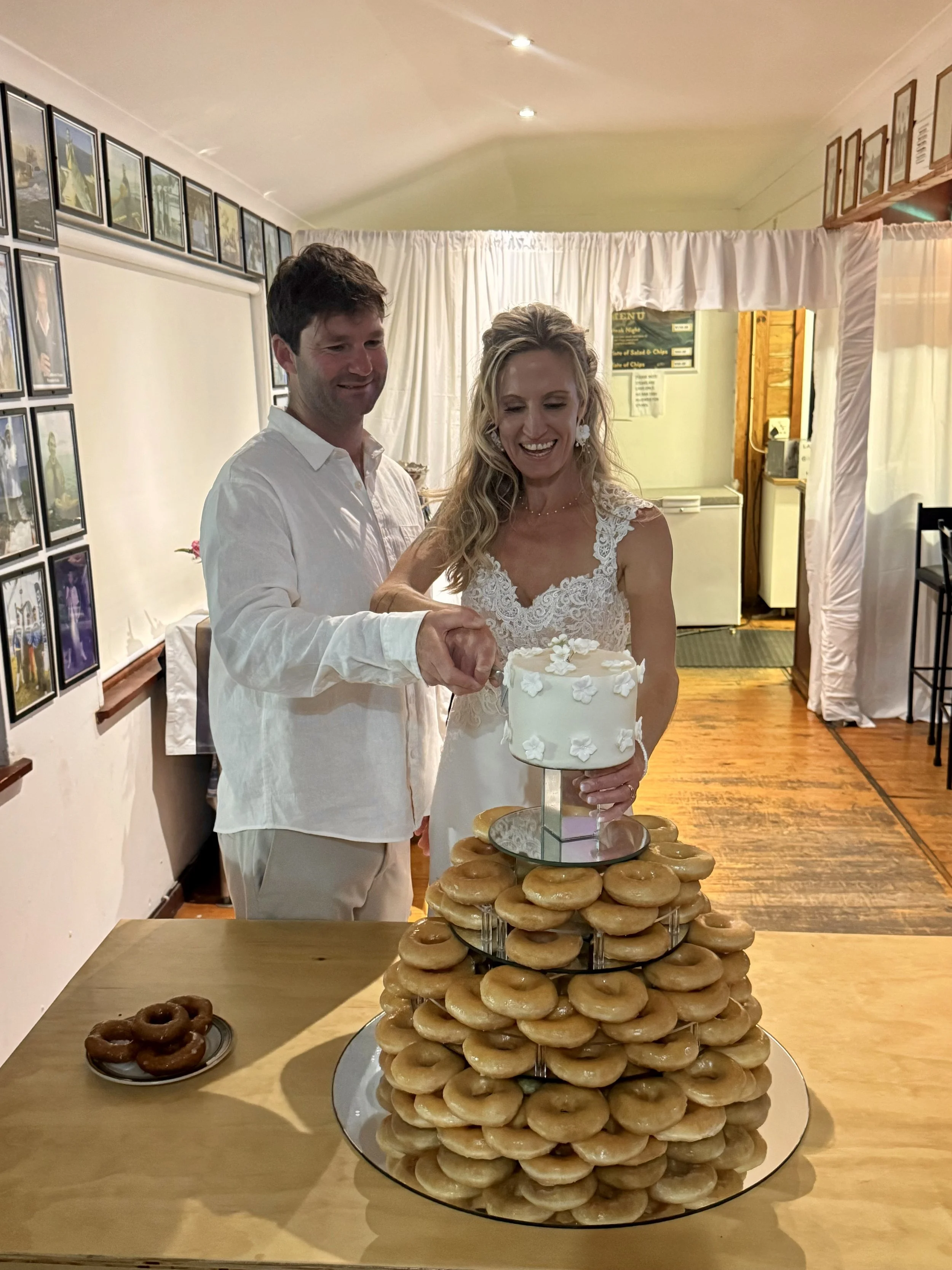 A bride and groom cutting a wedding cake together, surrounded by a large display of glazed donuts on a tiered stand at a wedding reception.