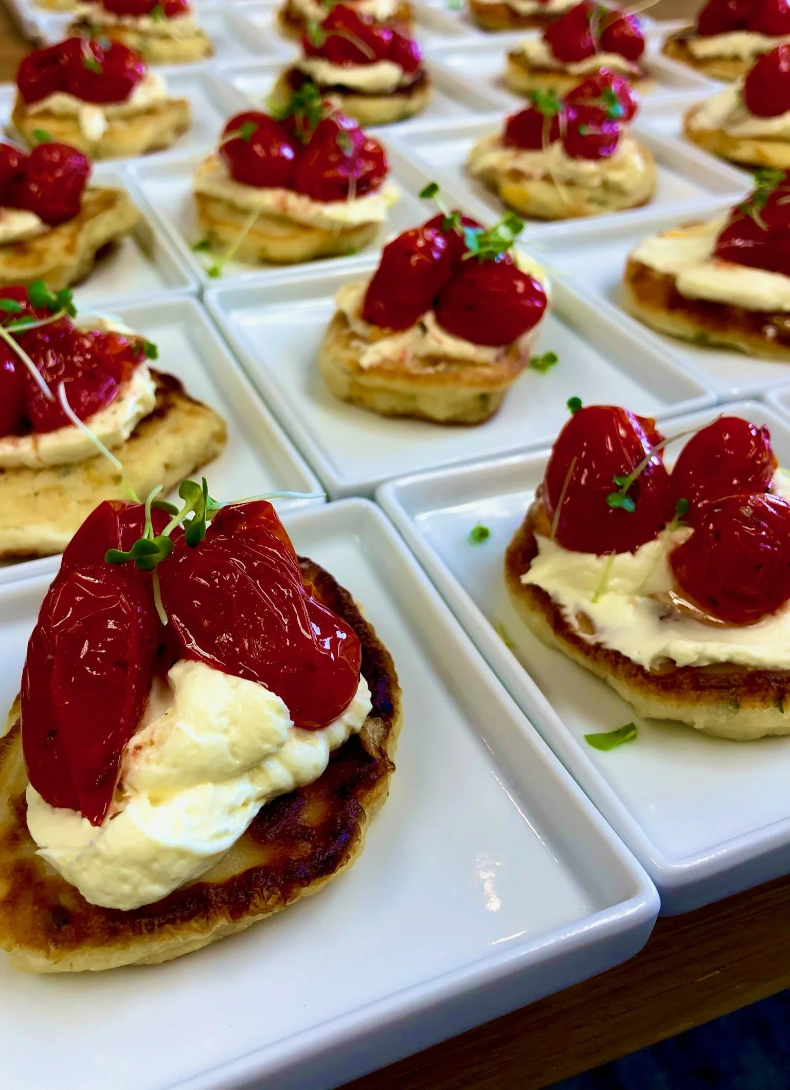 Appetizer servings of corn fritters topped with cheese, cherry tomatoes, and microgreens on small white plates.