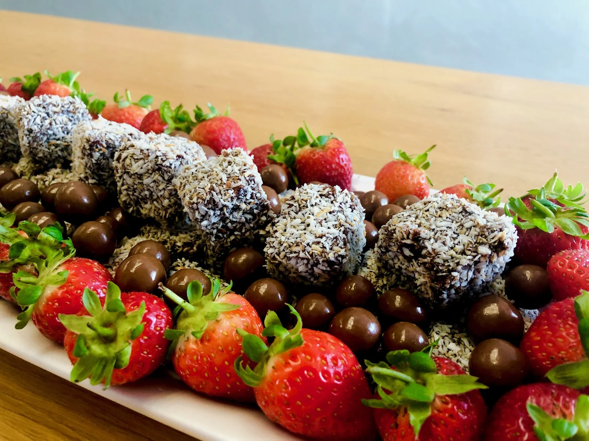 A white rectangular plate with fresh strawberries, chocolate-covered round treats, and chocolate candies arranged on a wooden table.