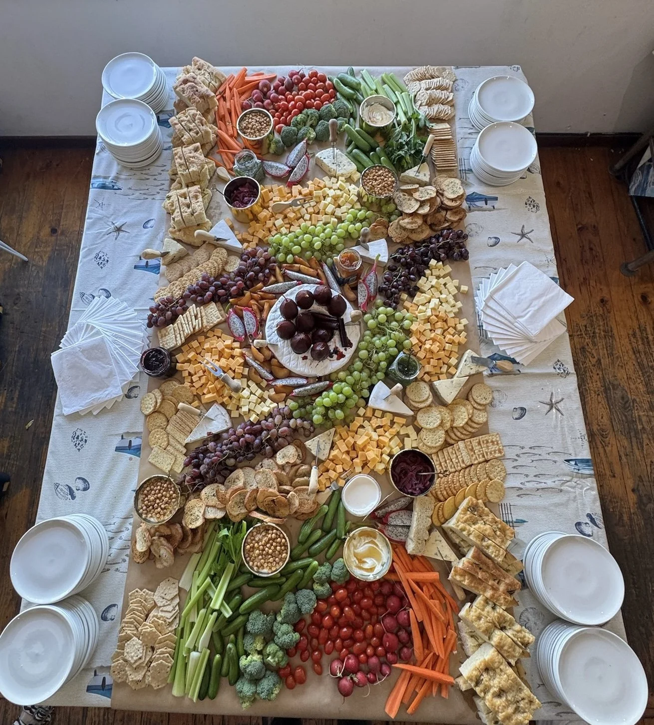 A table set with a large variety of cheeses, grapes, vegetables, and crackers, arranged in an elaborate platter for a gathering or celebration.