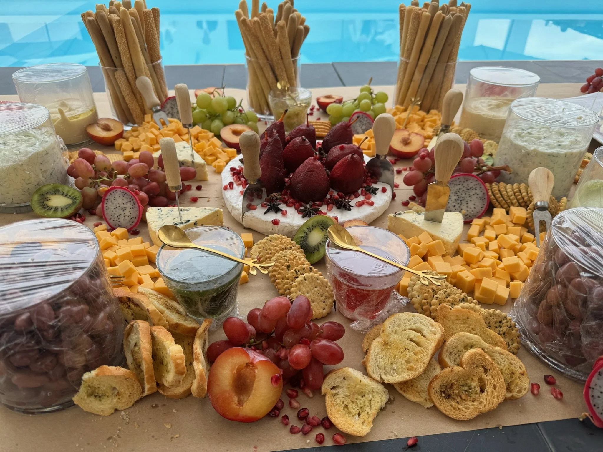 A cheese and fruit platter featuring various cheeses, grapes, strawberries, kiwi, pomegranate, crackers, bread, and a round cake topped with strawberries, cherries, and star anise, set on a table near a pool.