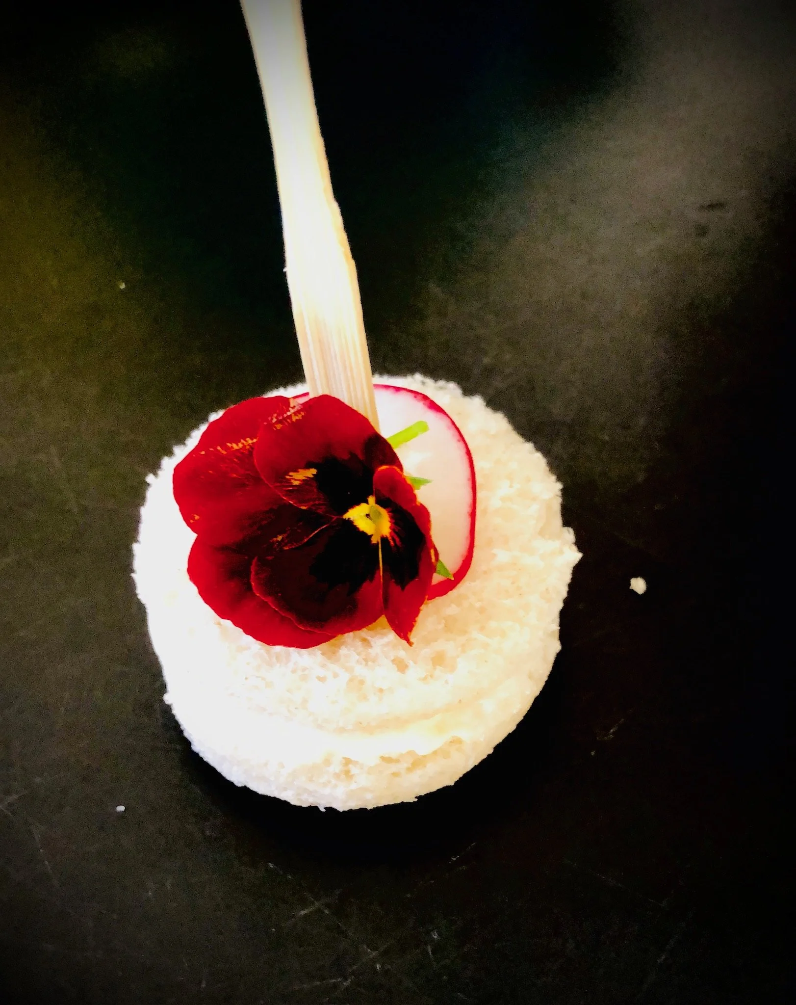 Round white dessert topped with a red and black edible flower and a small green garnish, with a wooden stick inserted upright.