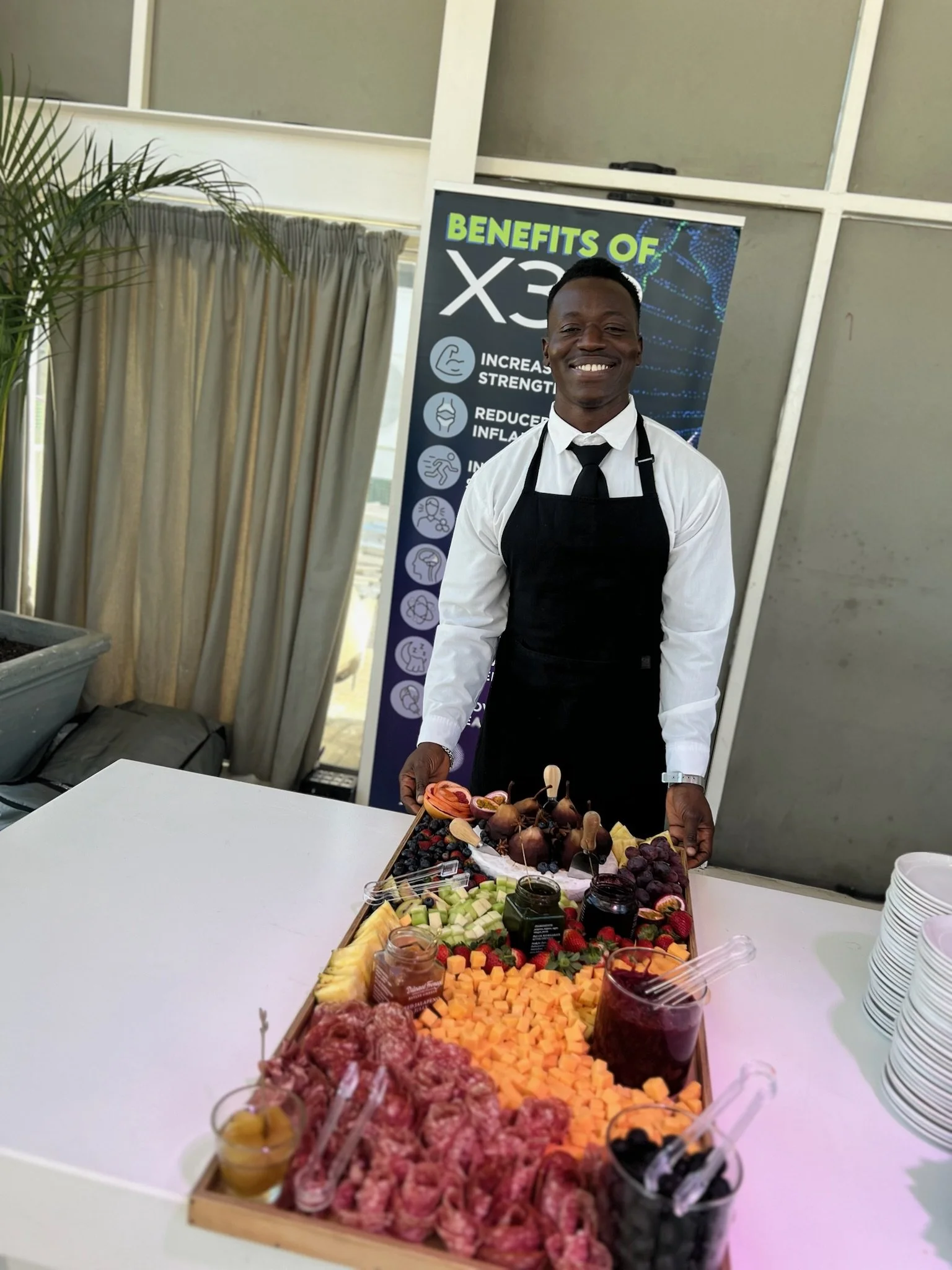 A man dressed in a white shirt, black tie, and black apron stands behind a table with a variety of fresh fruit and jam jars, smiling at the camera. Behind him is a sign about the benefits of X3.