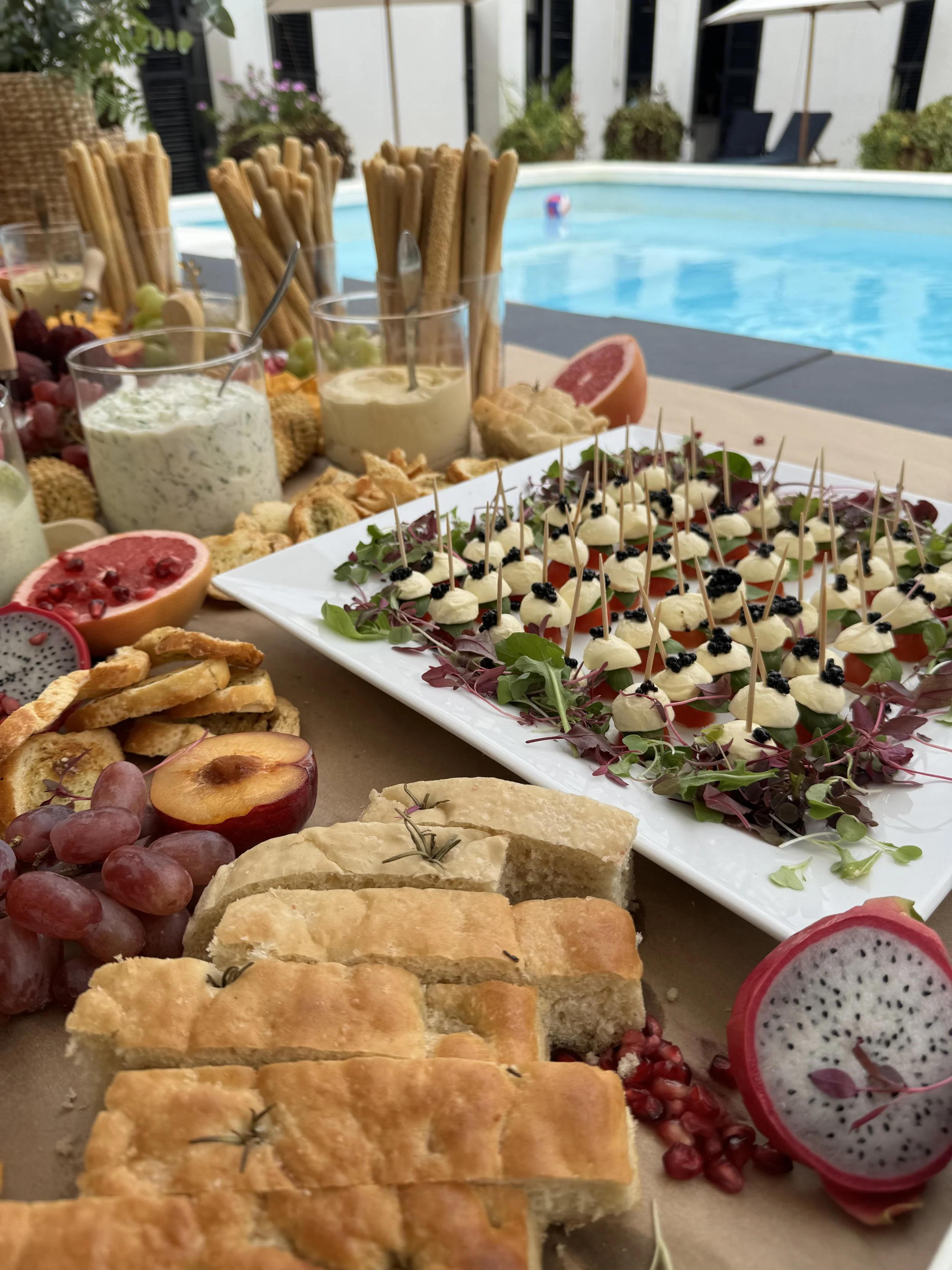 A poolside buffet with assorted appetizers and desserts, including bread, grapes, pomegranate, and a vegetable platter, with a swimming pool in the background.