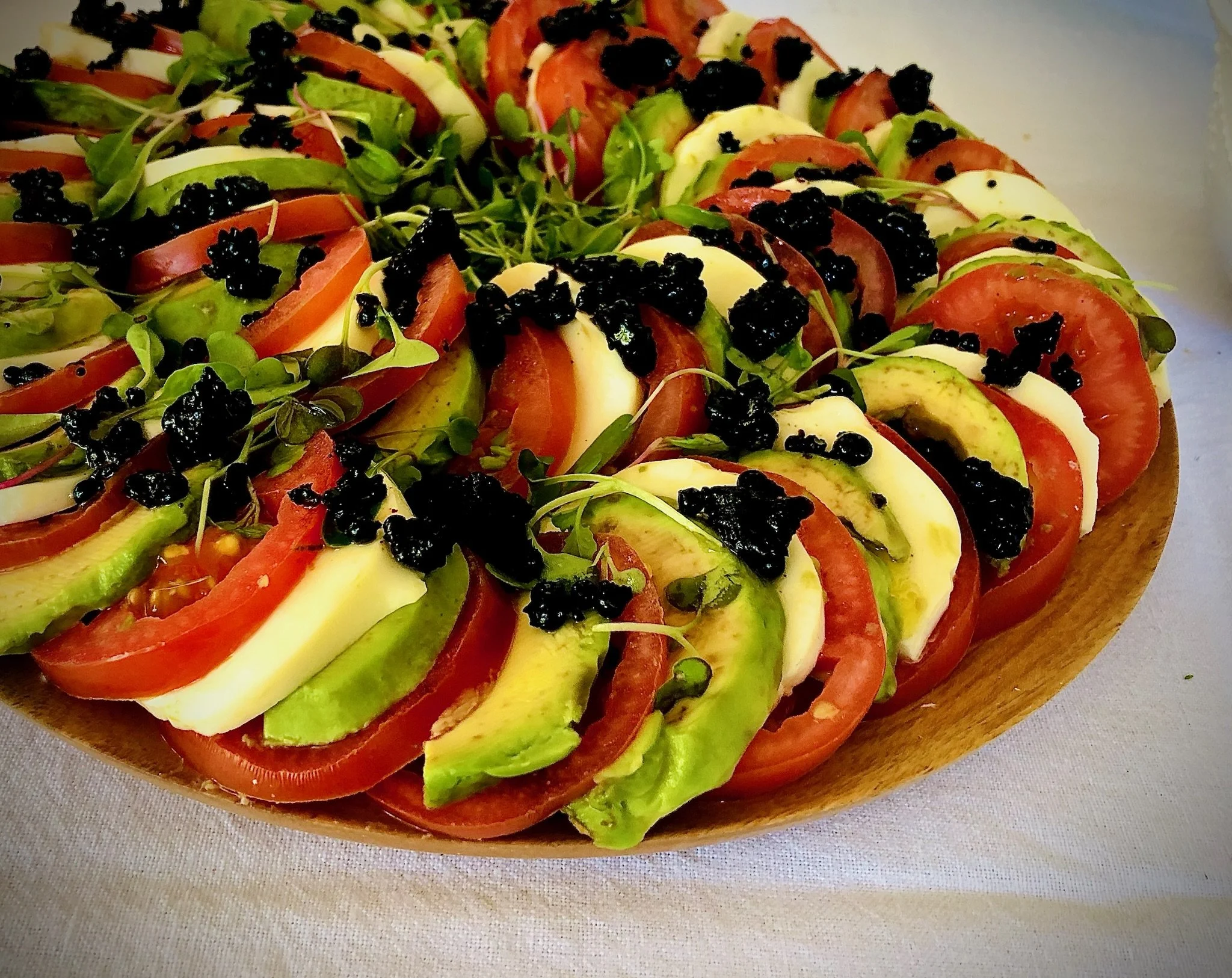 Caprese salad with sliced tomatoes, mozzarella cheese, avocado, green herbs, and black caviar on a wooden platter.