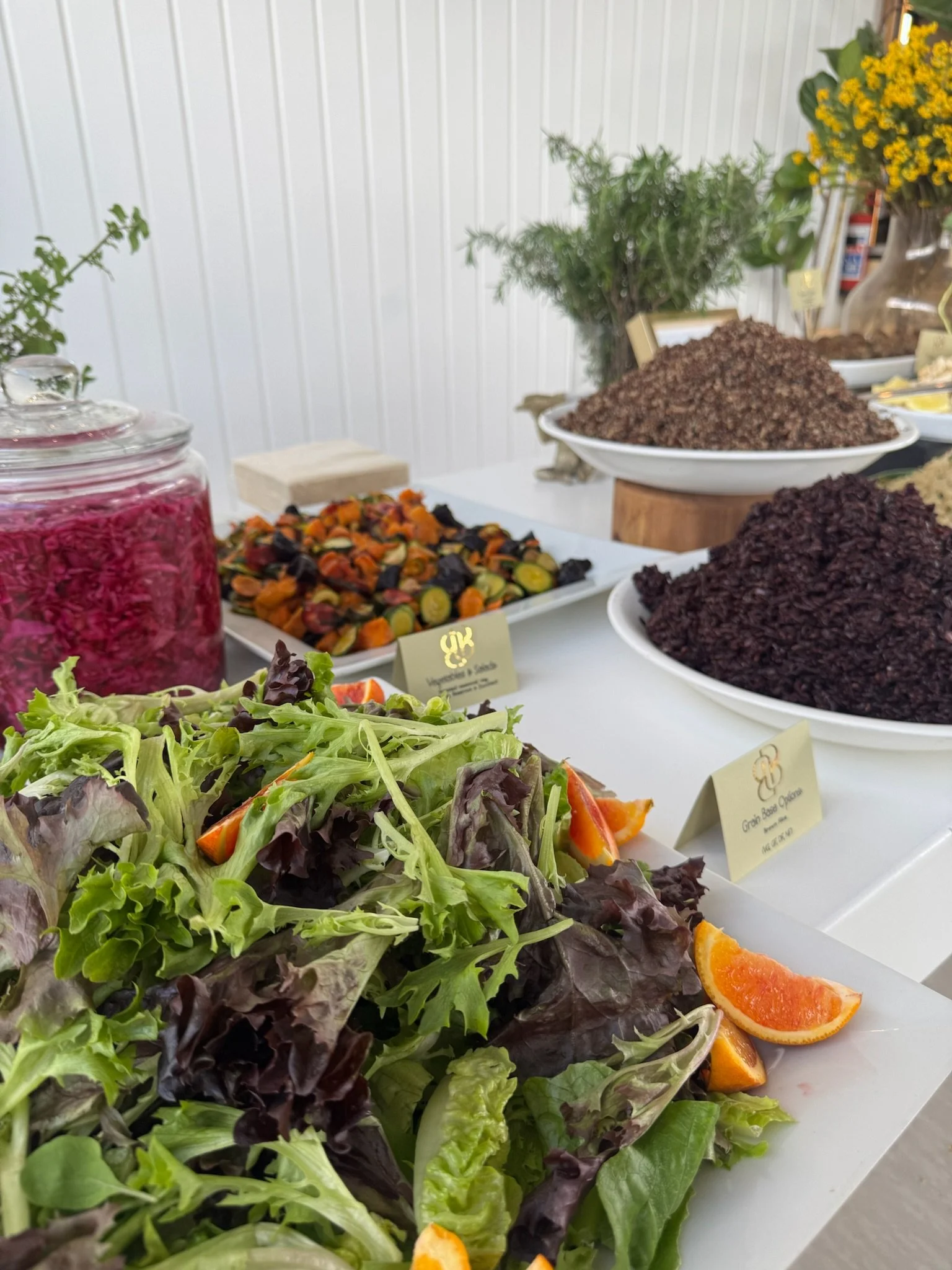 Fresh mixed green salad with orange wedges at a buffet table, with various bowls of grains and cooked vegetables in the background.