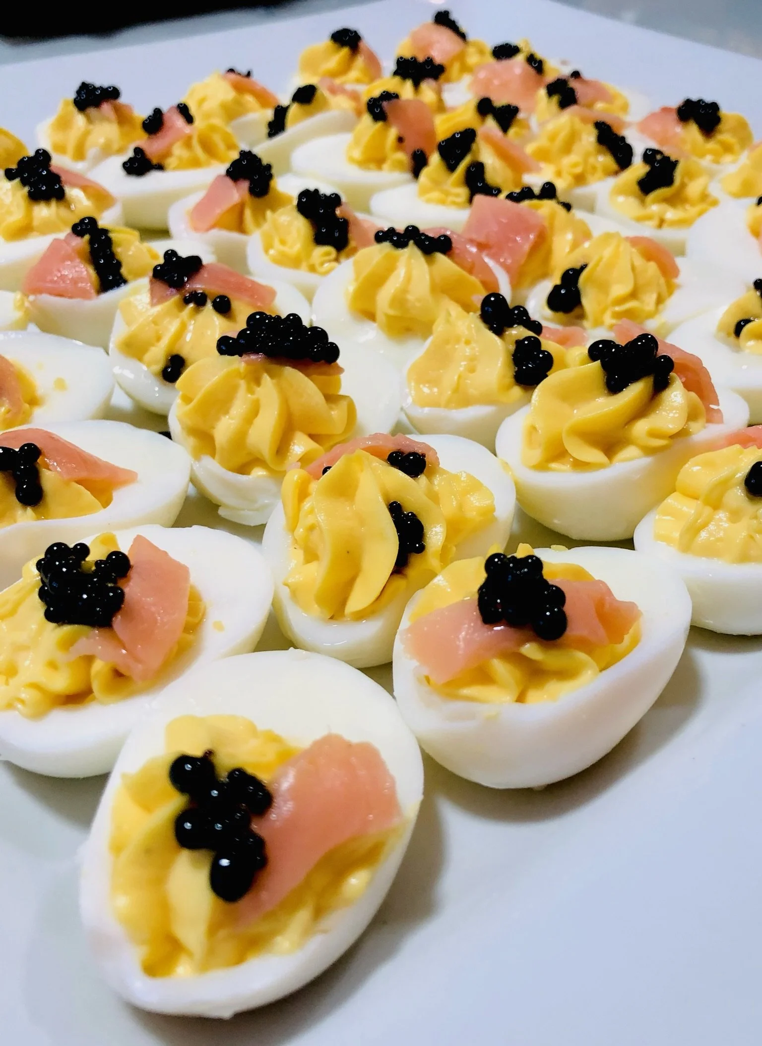 Close-up of deviled eggs topped with yellow yolk mixture, black caviar, and small slices of salmon, arranged on a white platter.