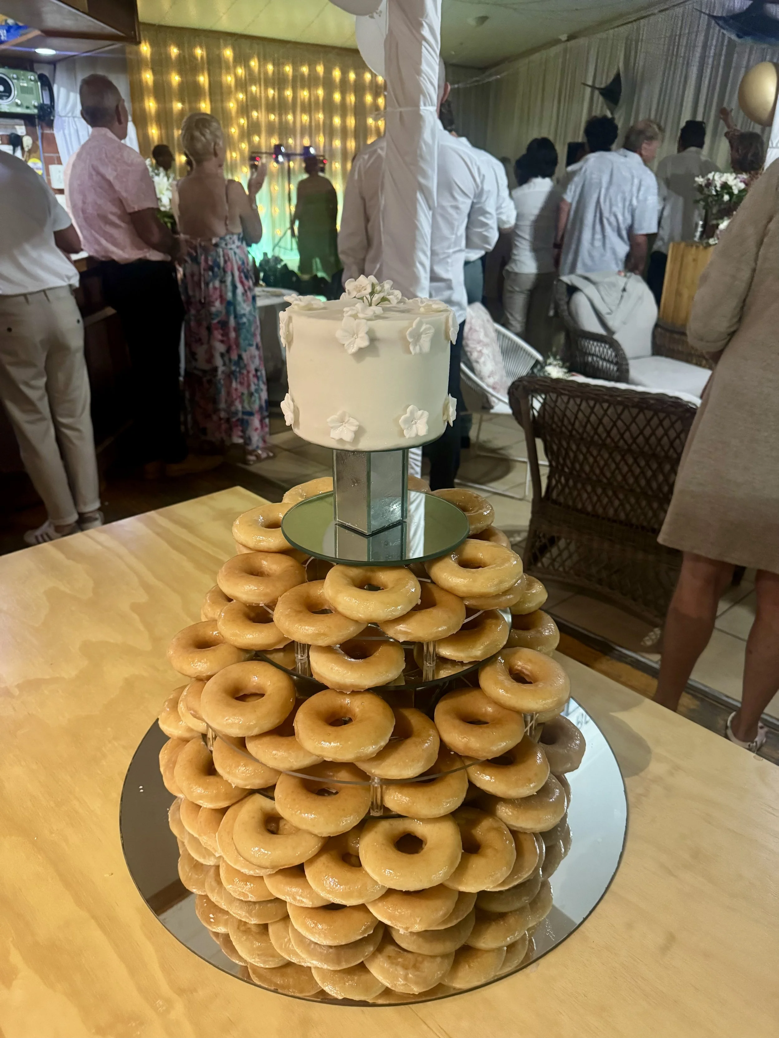 A tower of glazed donuts topped with a small white cake decorated with flowers, set upon a round mirror on a wooden table at a celebration or party.