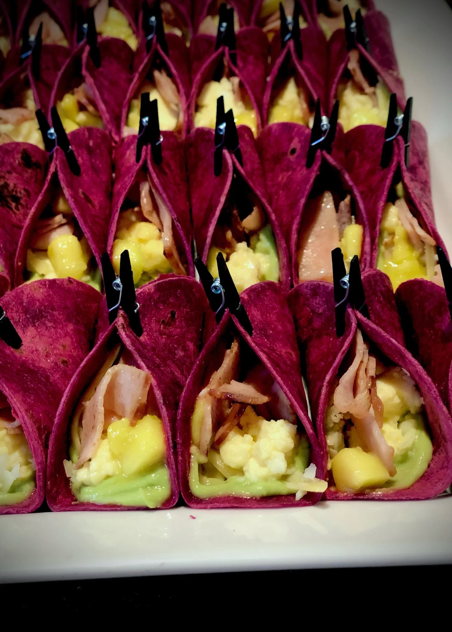 Close-up of small wraps with purple beetroot tortillas, filled with cheese, shredded meat, and vegetables, secured with black toothpicks.