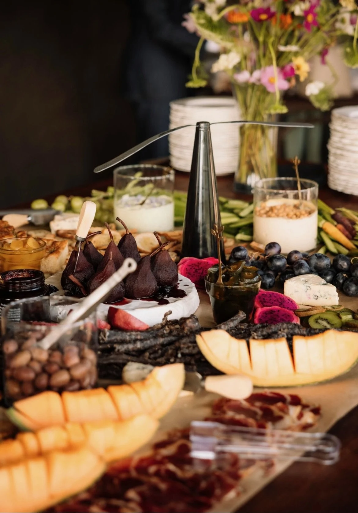 A spread of various cheeses, fresh fruits, and snacks on a wooden table, with a bouquet of flowers and stacked white plates in the background.