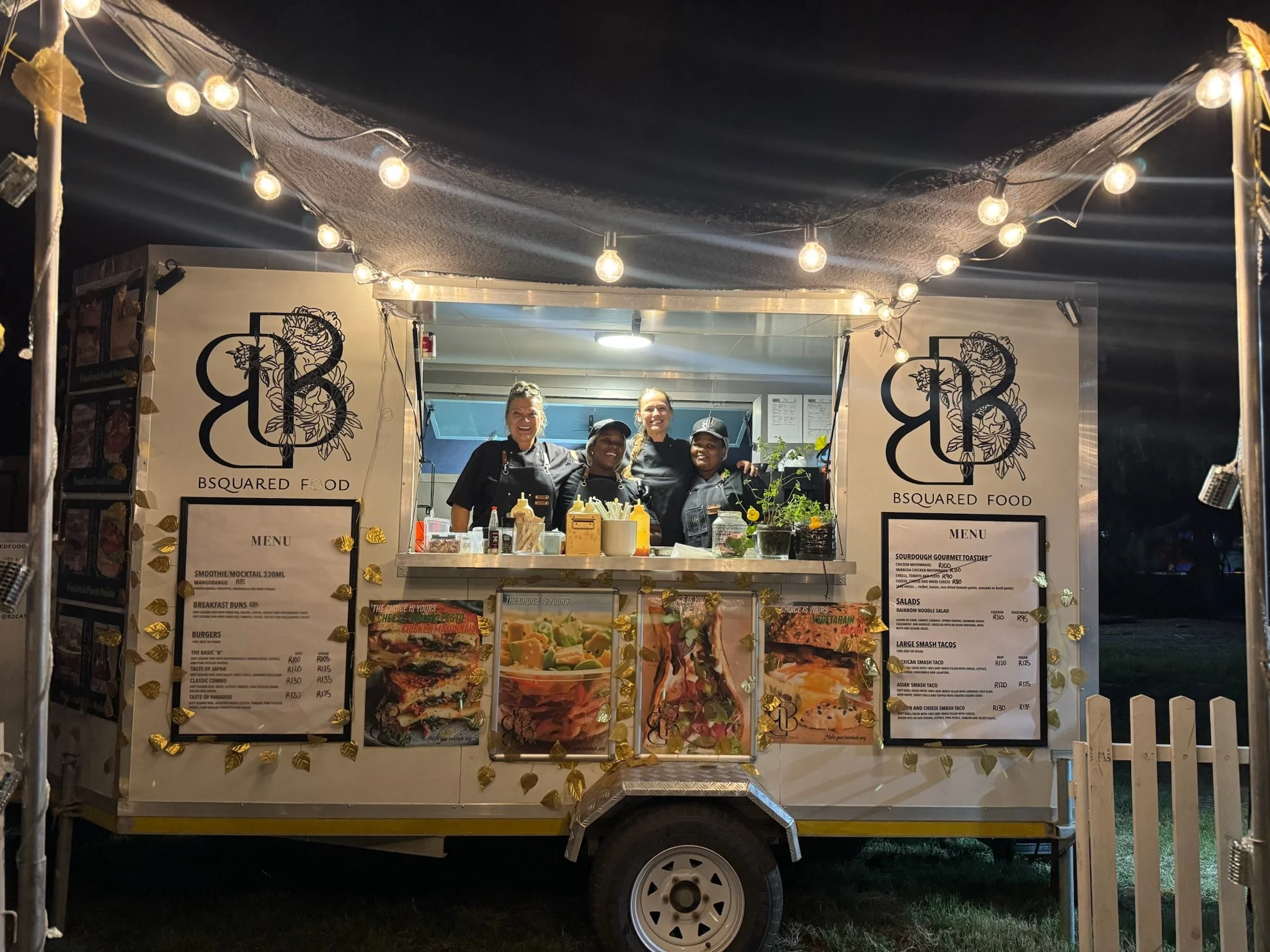 Food truck named 'B Squared Food' with four smiling staff members inside, illuminated by string lights, at night, with menus displayed on the sides and a picket fence at the front.