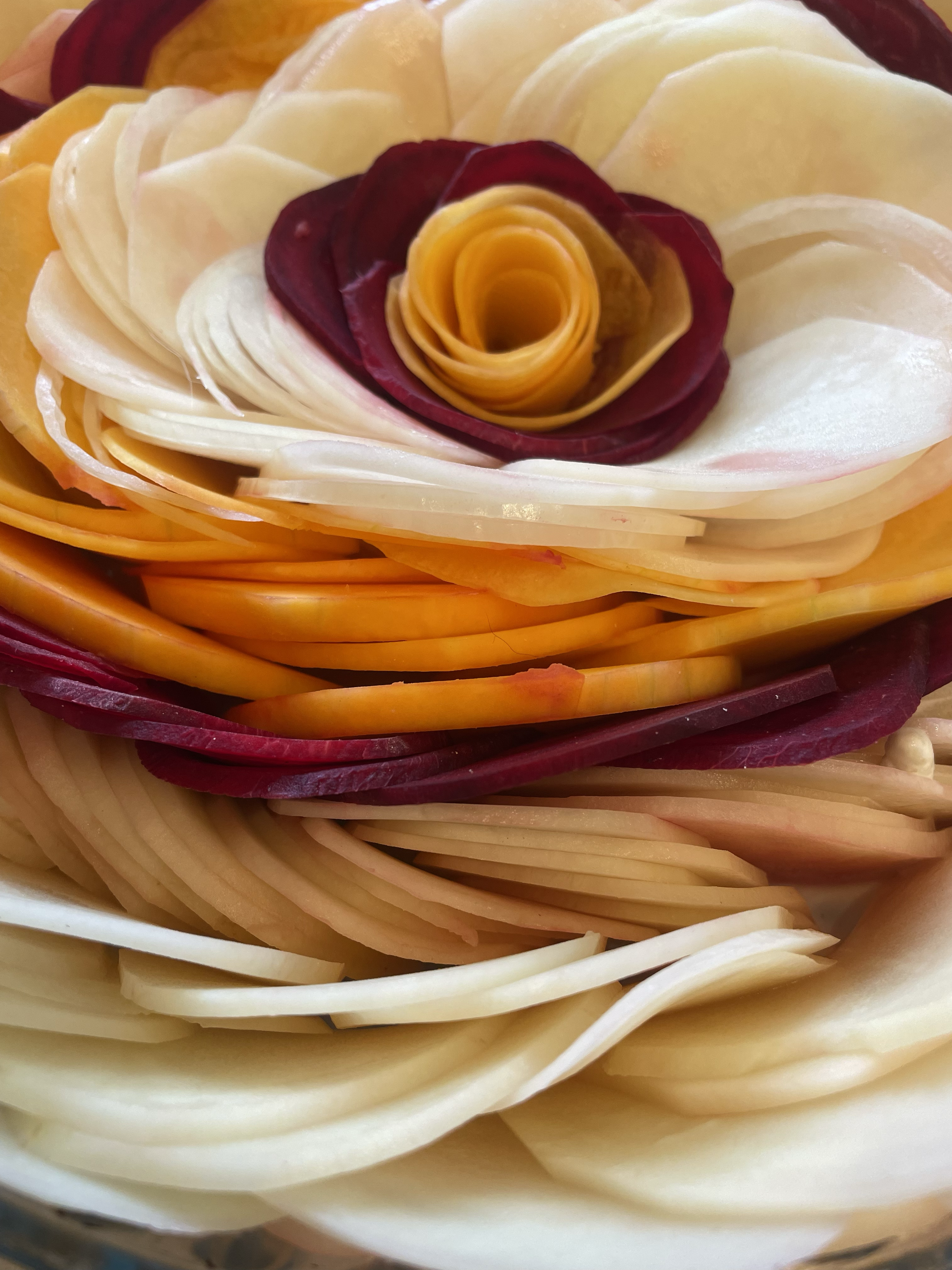 Close-up of thinly sliced vegetables arranged in a layered, flower-like pattern, with a spiral of yellow and red slices in the center.