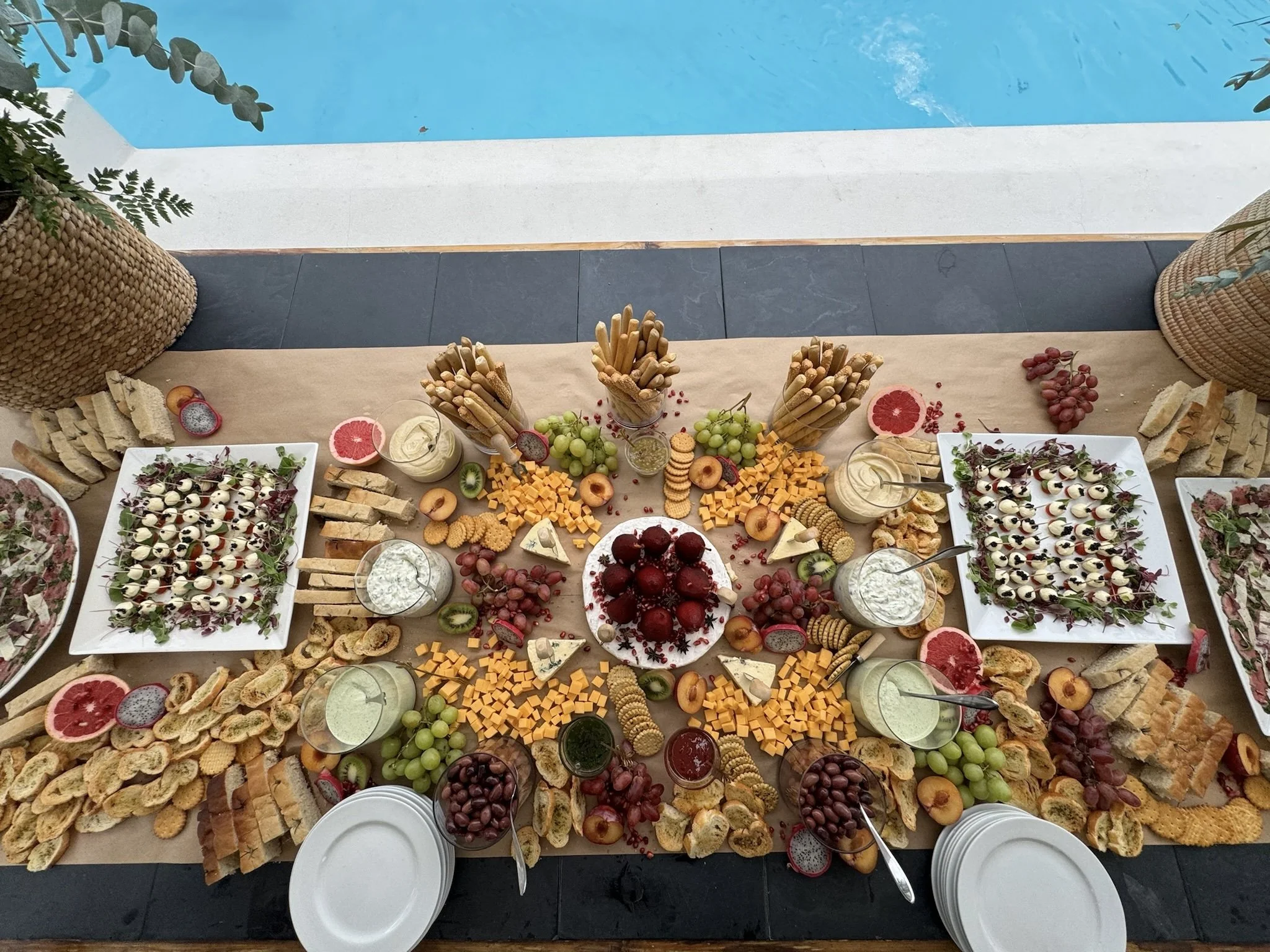 A large table with an assortment of cheeses, fruits, crackers, bread, salad, and dips, arranged for a meal or party, with a view of a pool in the background.