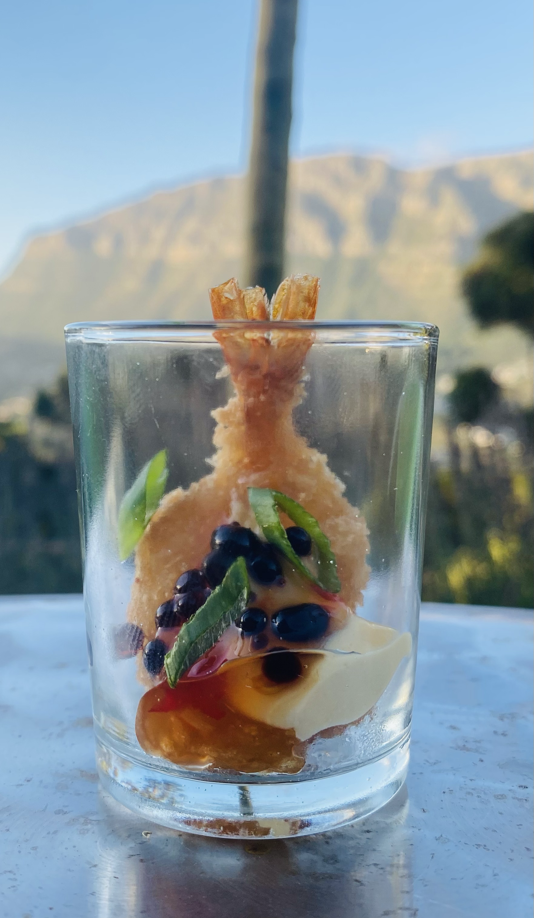 Close-up of a glass containing a prawn cocktail with a lemon wedge, black caviar, and green herbs, with a mountain and clear sky background.