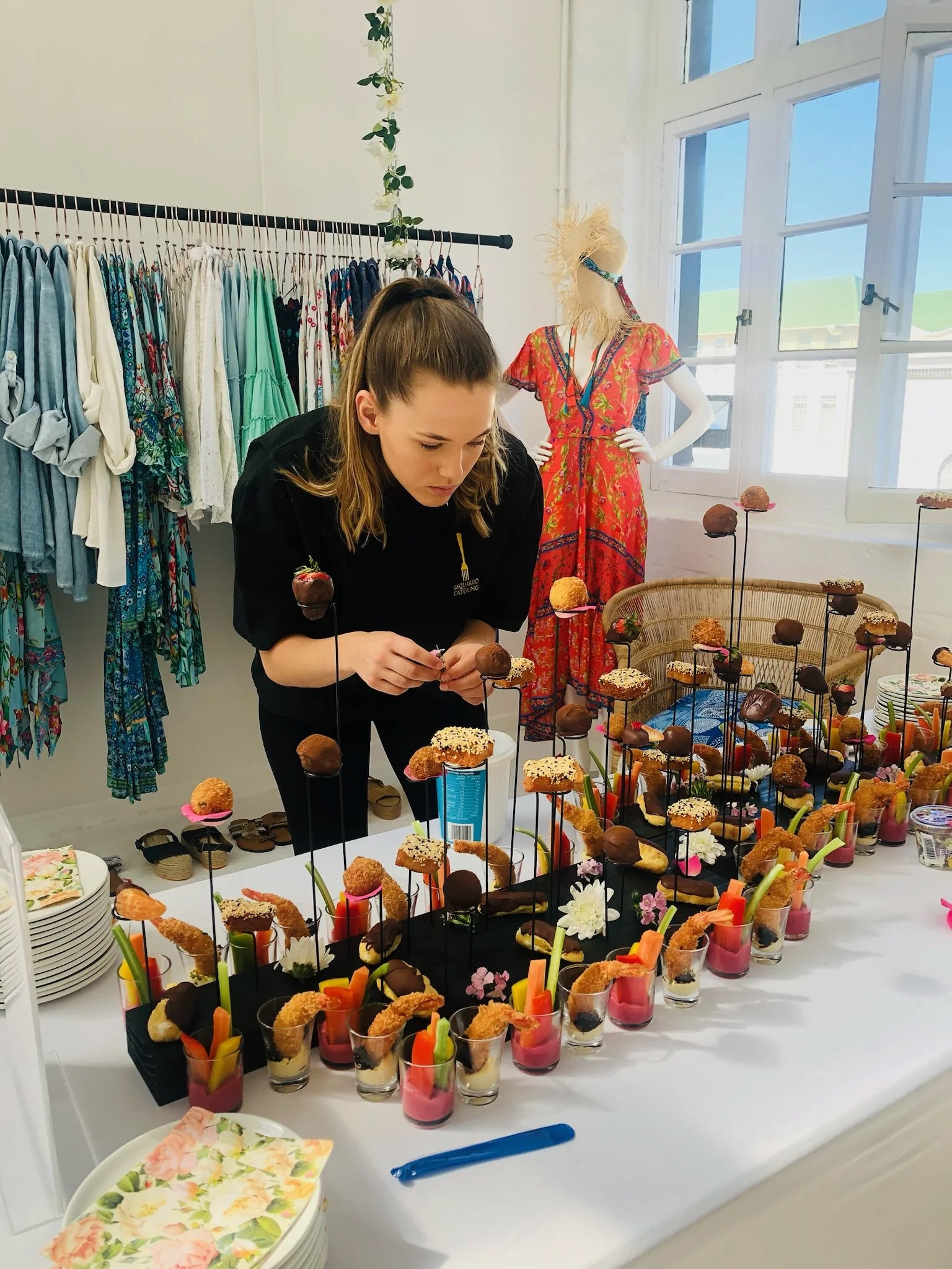 A young woman arranging appetizers or desserts on a table at a party or event, with colorful appetizers arranged in small cups in front of her and a mannequin dressed in a bright red dress in the background.