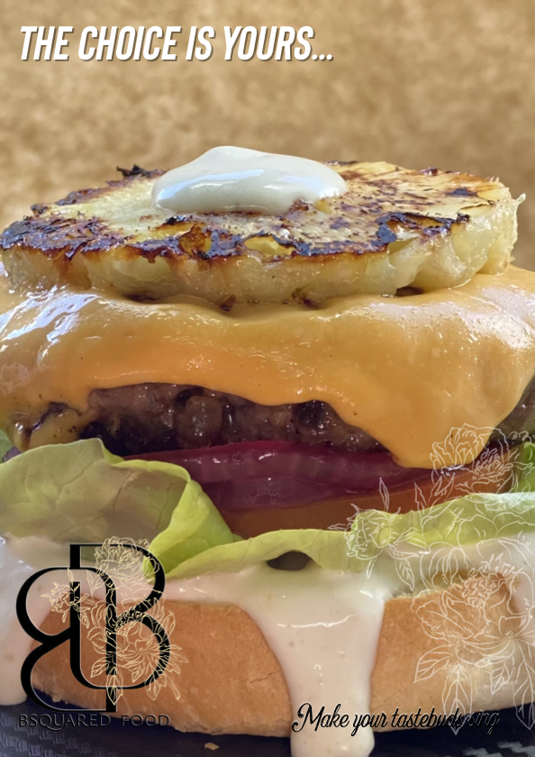 Close-up of a layered cheeseburger with lettuce, tomato, melted cheddar cheese, a beef patty, a grilled pineapple slice, and mayonnaise on a bun, with the text "The choice is yours..." and "Make your taste buds sing."