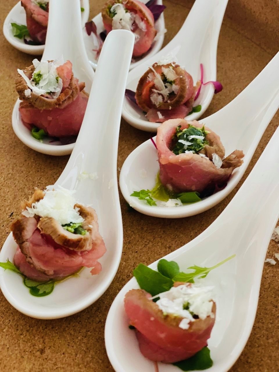 Appetizer bites served on white ceramic spoons, featuring rolled slices of beef with cheese and herbs, garnished with greens.