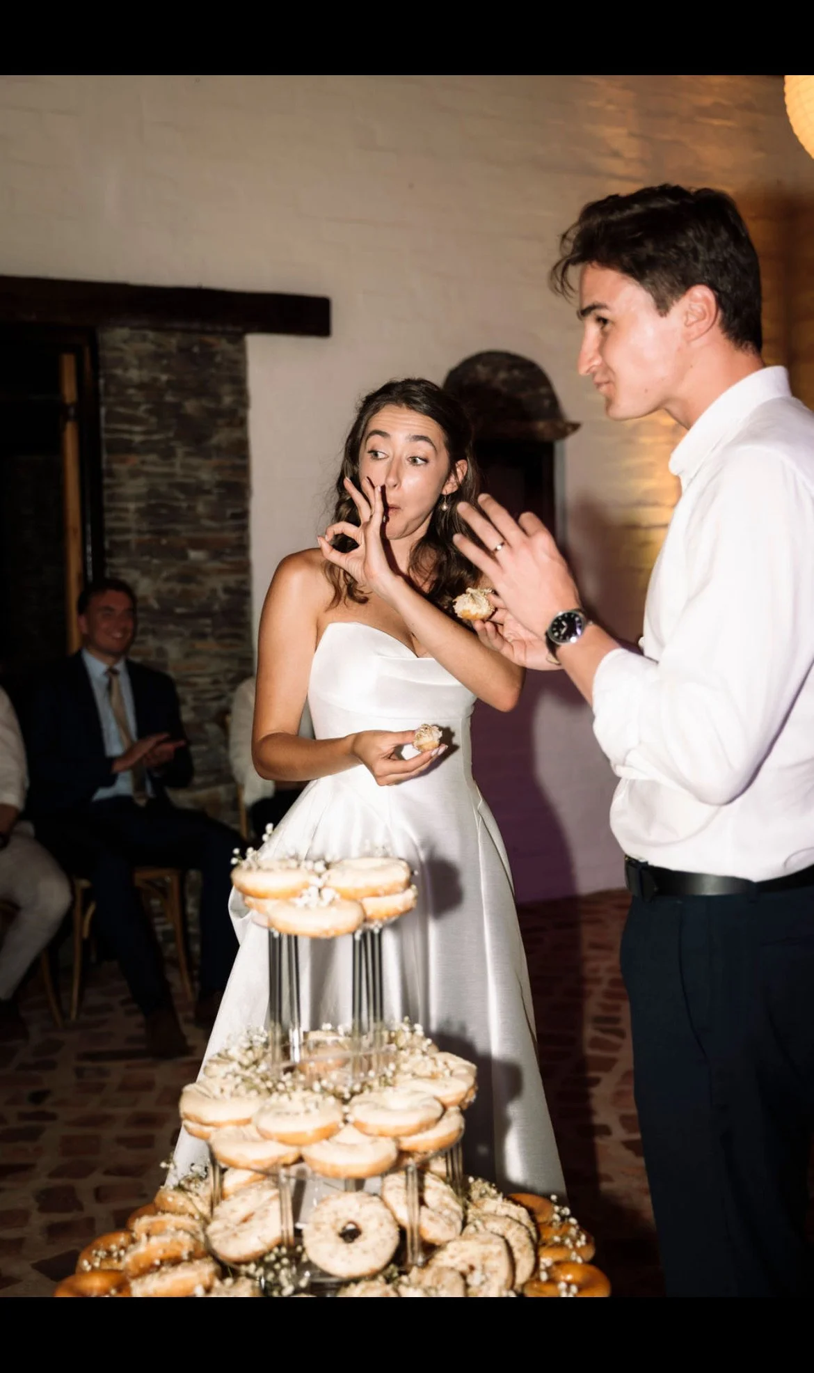 A bride and groom at a wedding reception with a tower of donuts in the foreground. The bride is wearing a white strapless wedding gown and is making a gesture with her finger to her lips, while the groom is dressed in a white shirt and dark pants, ho