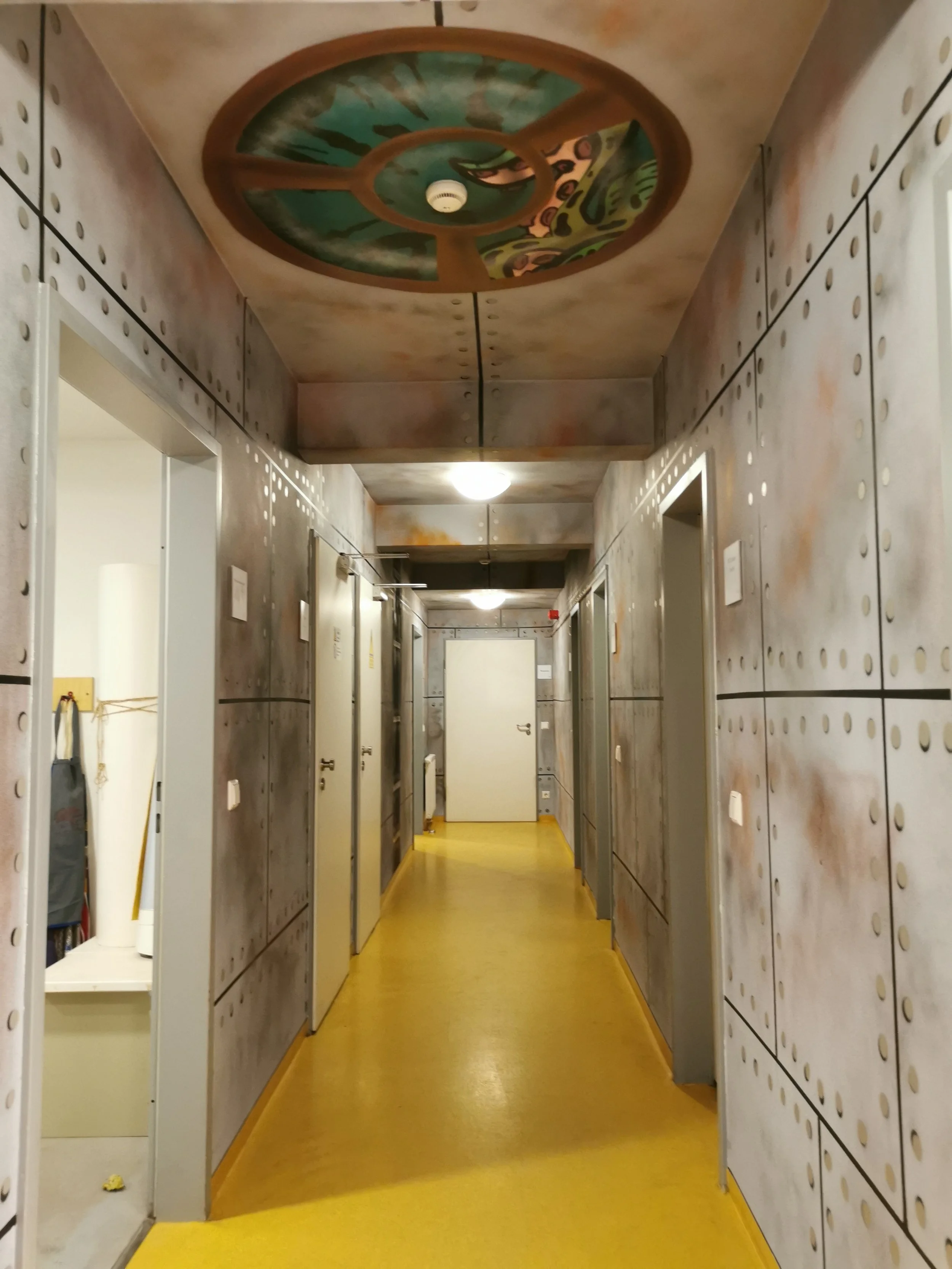 Ein Flur in einem Gebäude mit gelber Fußboden, grauen Wänden und mehreren Türen. Die Decke ist mit einem kunstvollen, bemalten Rundpanel gestaltet.