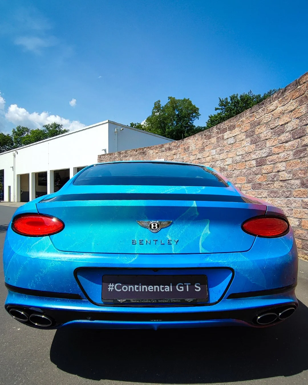 Blaue Bentley Continental GT S mit vier Auspuffrohren, parkend vor einer Steinmauer