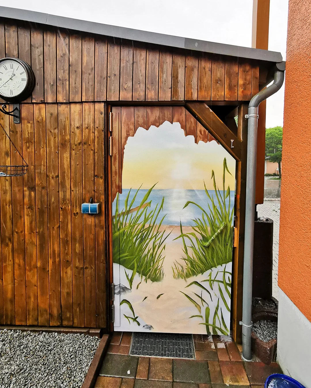 Eine Tür mit einem handgemalten Strandbild, das den Blick aufs Meer, Sand und Gras zeigt, umgeben von einer Holzfassade.