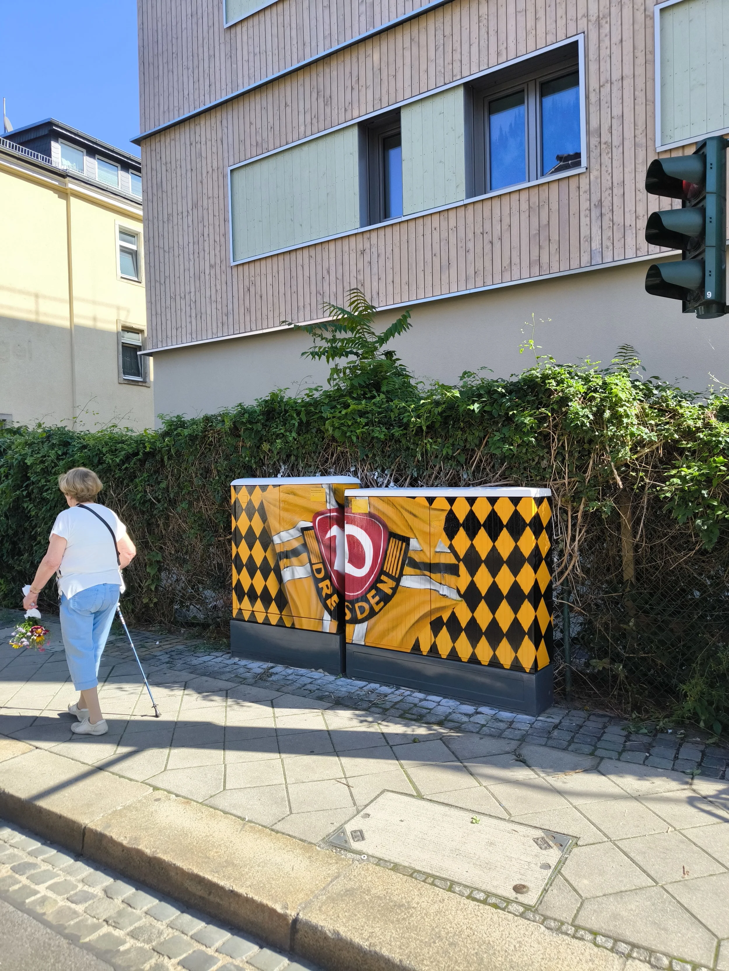 Eine ältere Frau mit weißen Haaren, weißen T-Shirt, blauen Hosen und Rollator, die Blumen hält, steht auf einem Bürgersteig neben einer mit einem Borussia Dortmund Logo bemalten elektrischen Verteilerboxen. Im Hintergrund sind moderne Gebäude und ein