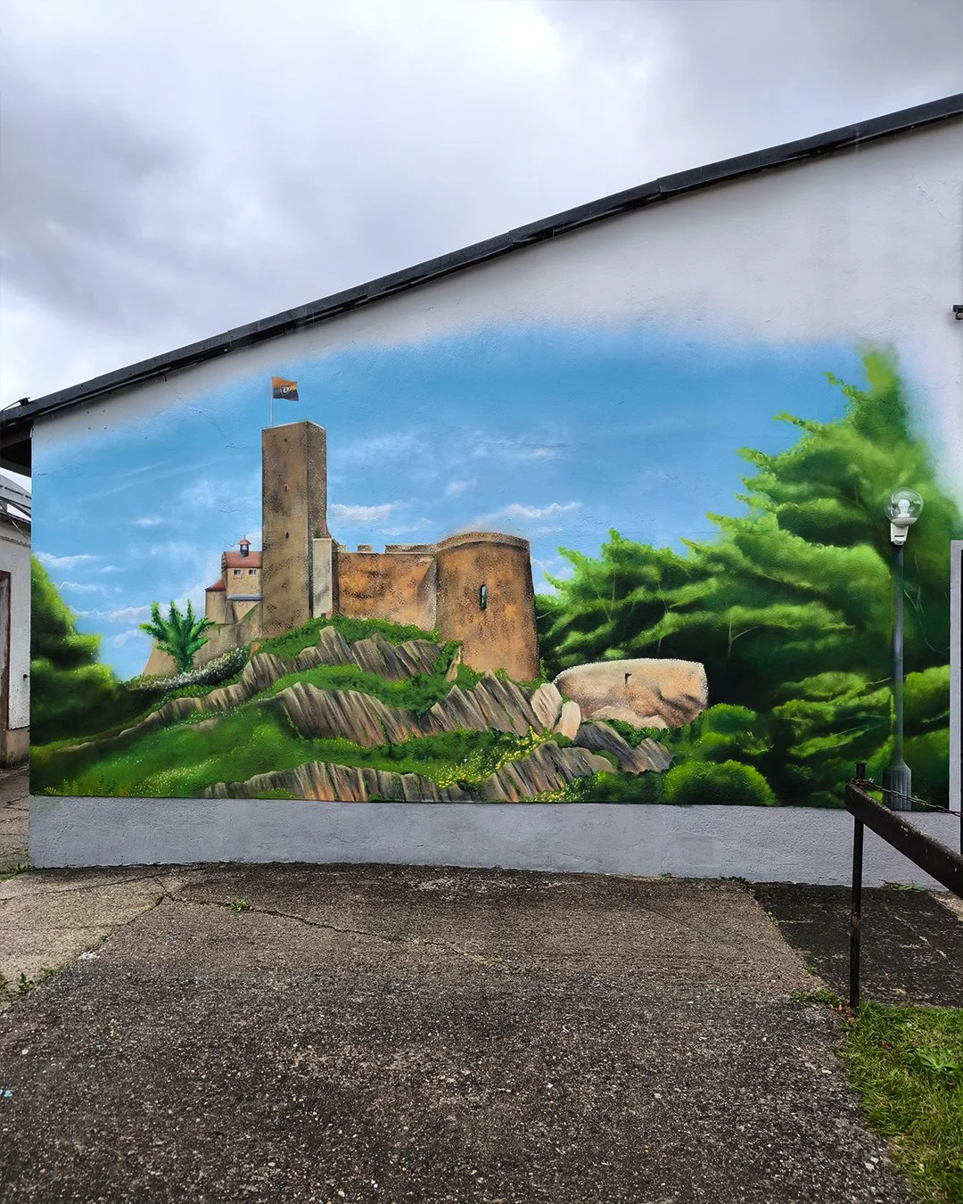 Straßenmauer mit Wandgemälde eines Schlosses auf einem Hügel, umgeben von grünen Bäumen und Felsen. Das Gemälde zeigt einen blauen Himmel mit Wolken.