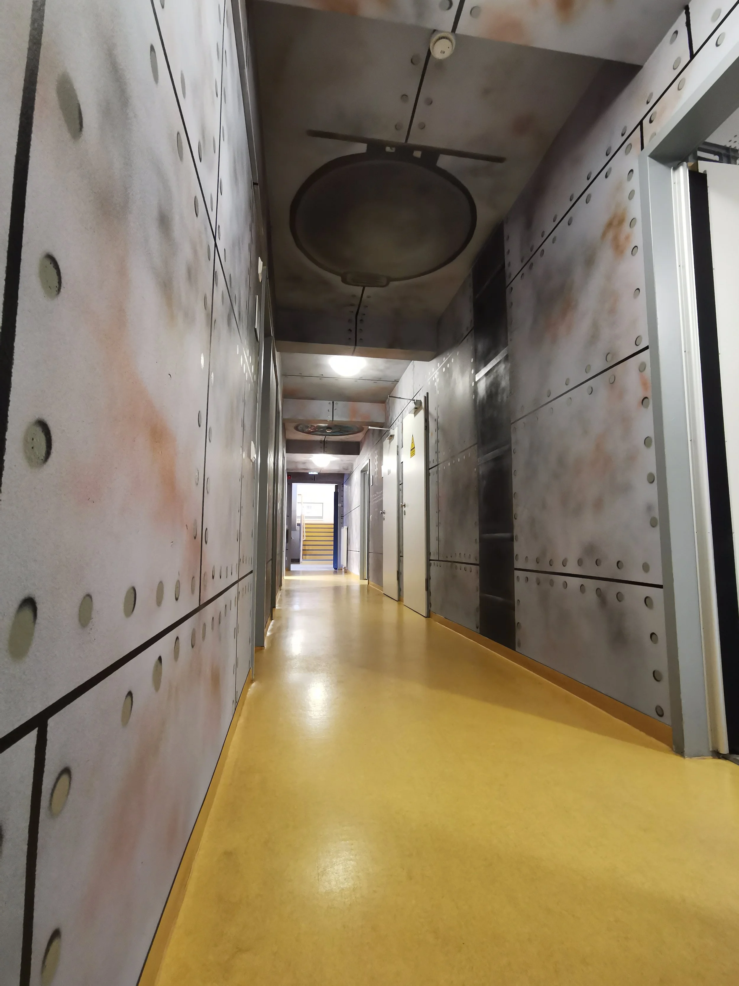 Langer Flur in einem Gebäude mit metallisch wirkenden Wänden und einem gelben Boden, Deckenventilatoren und Lichtleuchten