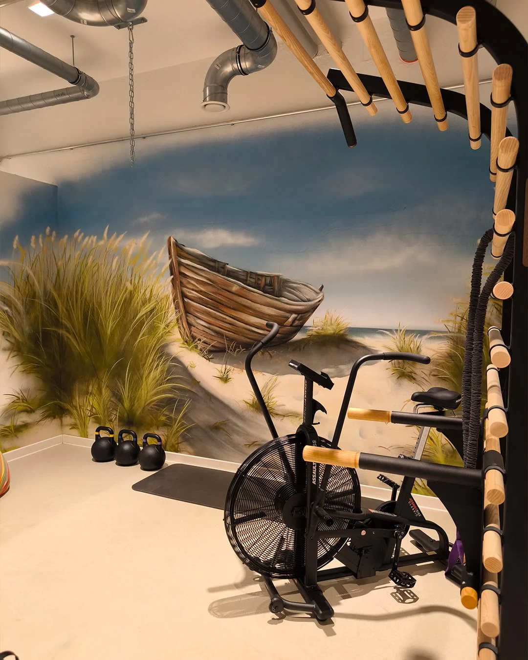 Fitnessraum mit Wandmalerei eines verrosteten Boots am Strand, drei schwarze Kettlebells, ein schwarzes Laufband, ein Fahrrad und eine Kletterwand mit Holzgriffen.