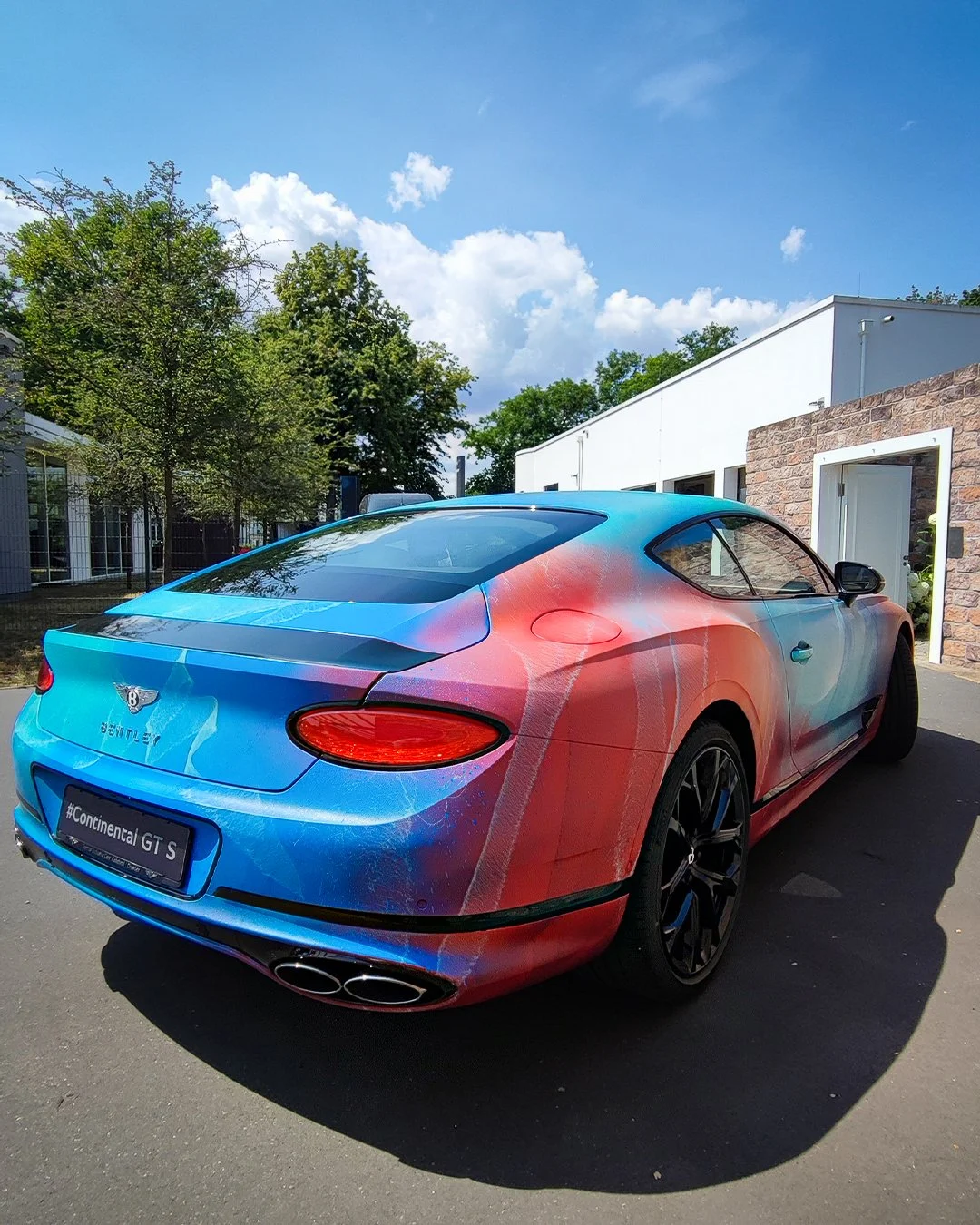 Ein sportlicher Bentley Continental GT S mit bunter Foliengestaltung, geparkt vor einem modernen Gebäude, bei sonnigem Himmel.