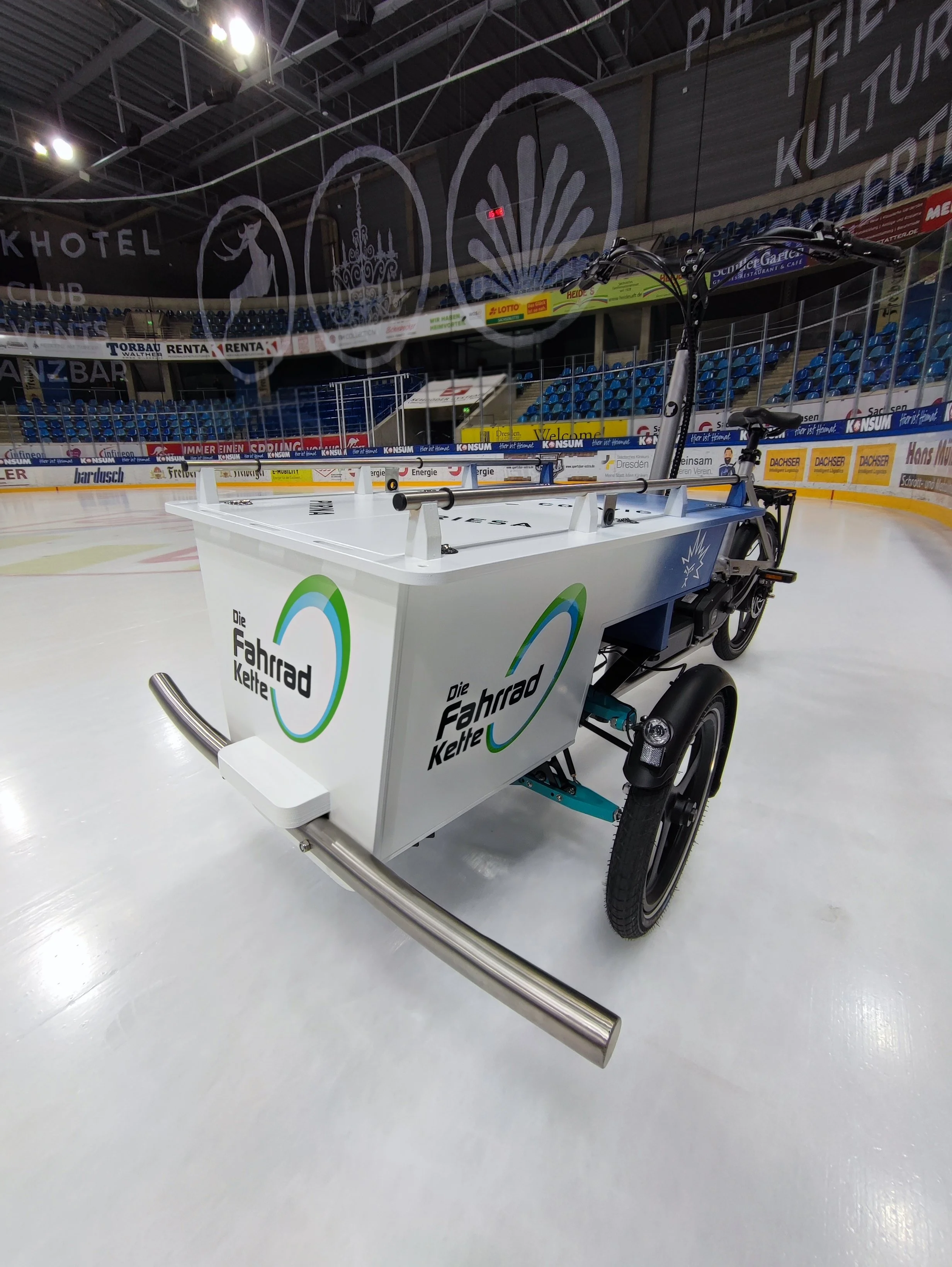 Ein Eisbahn mit einem E-Fahrrad, das auf das Eis gestellt ist, mit Werbung der "Die Fahrrad Kette" auf der Seite. Im Hintergrund sind blaue Sitzränge und Werbebanner sichtbar.
