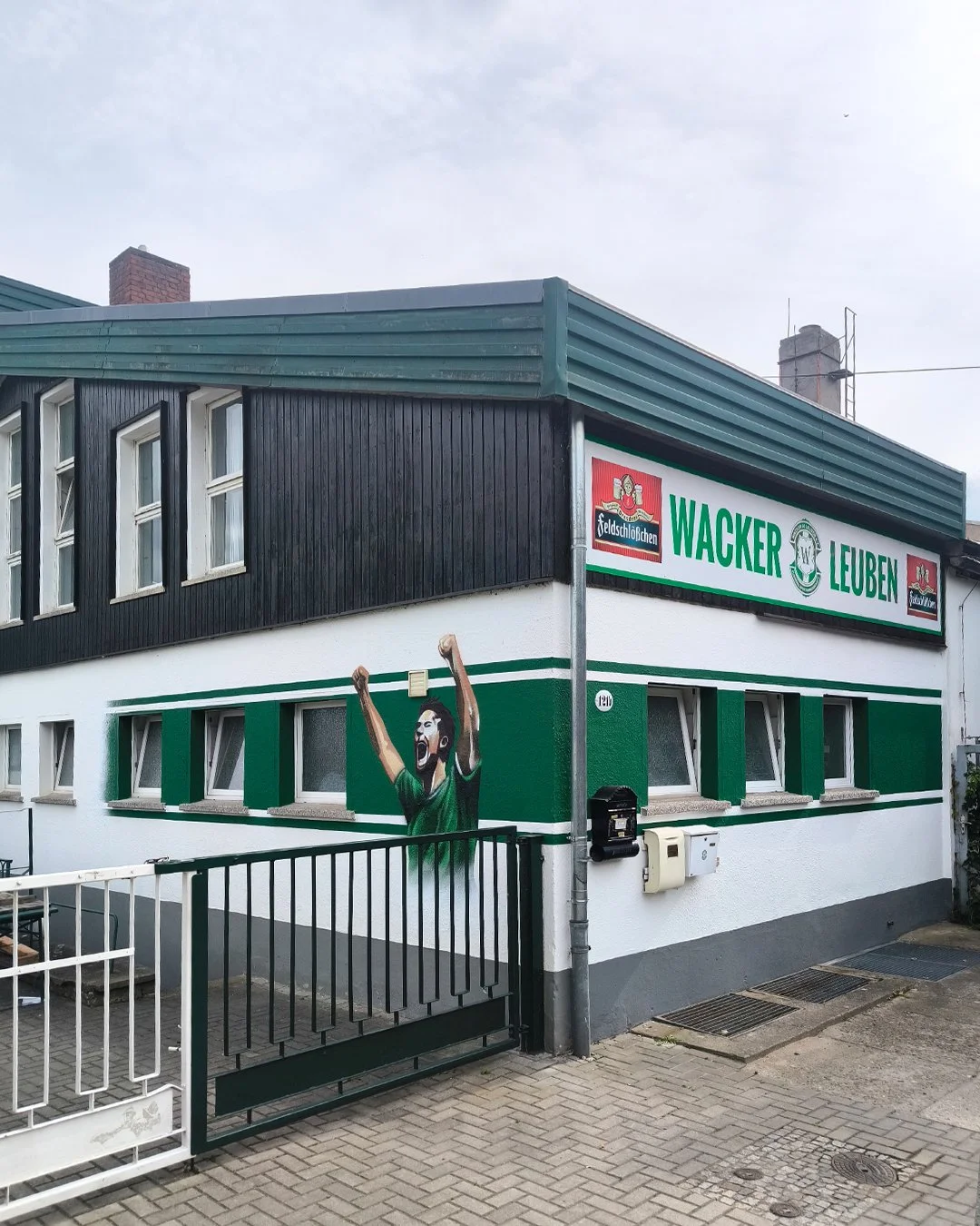Straßenszene mit einem Haus, das an einer Ecke eine Wandmalerei eines jubelnden Mannes zeigt, und einem Werbeschild der Brauerei Wacker Leuben.