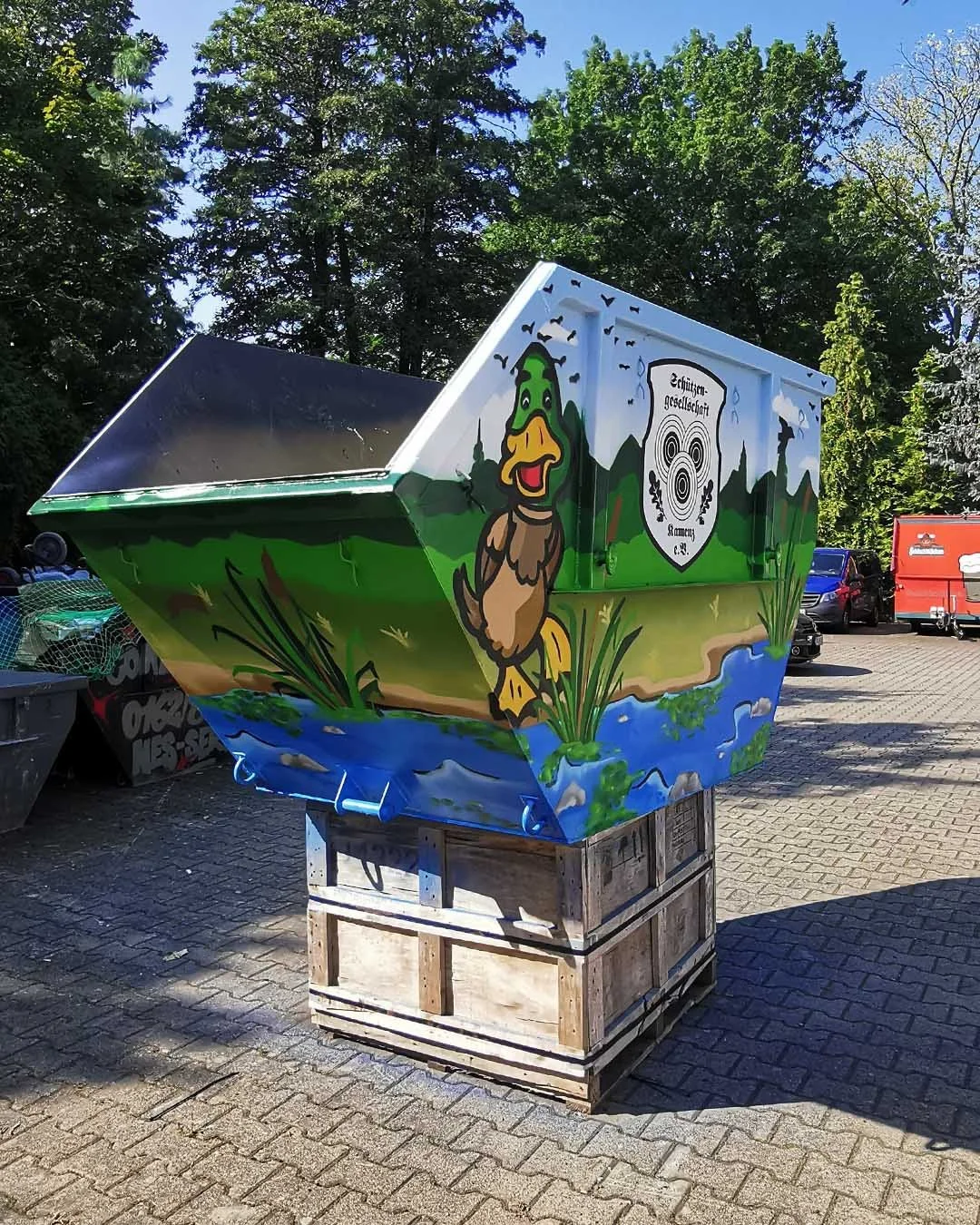 Ein bunter, handbemalter Braunkohletank mit einem Maskottchen eines fröhlichen Erpels und dem Wappen der Schützen-Gesellschaft. Der Tank steht auf einer Holzpalette auf einem gepflasterten Platz, im Hintergrund sind Autos und grüne Bäume.
